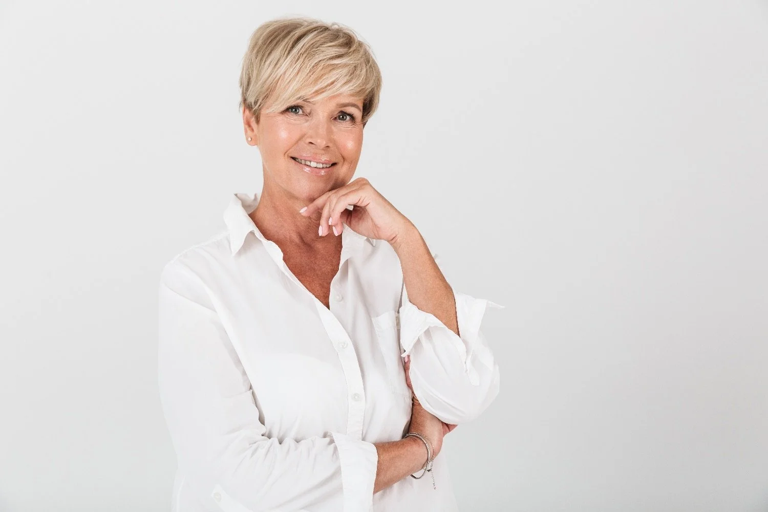 Female with short blonde hair wearing a white shirt smiles, holding her chin lightly in one of her hands.