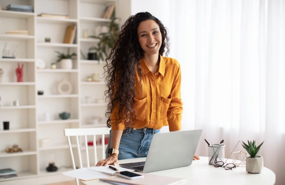 Happy middle eastern female entrepreneur standing near a laptop at a desk - she wears a bright mustard colour shirt.