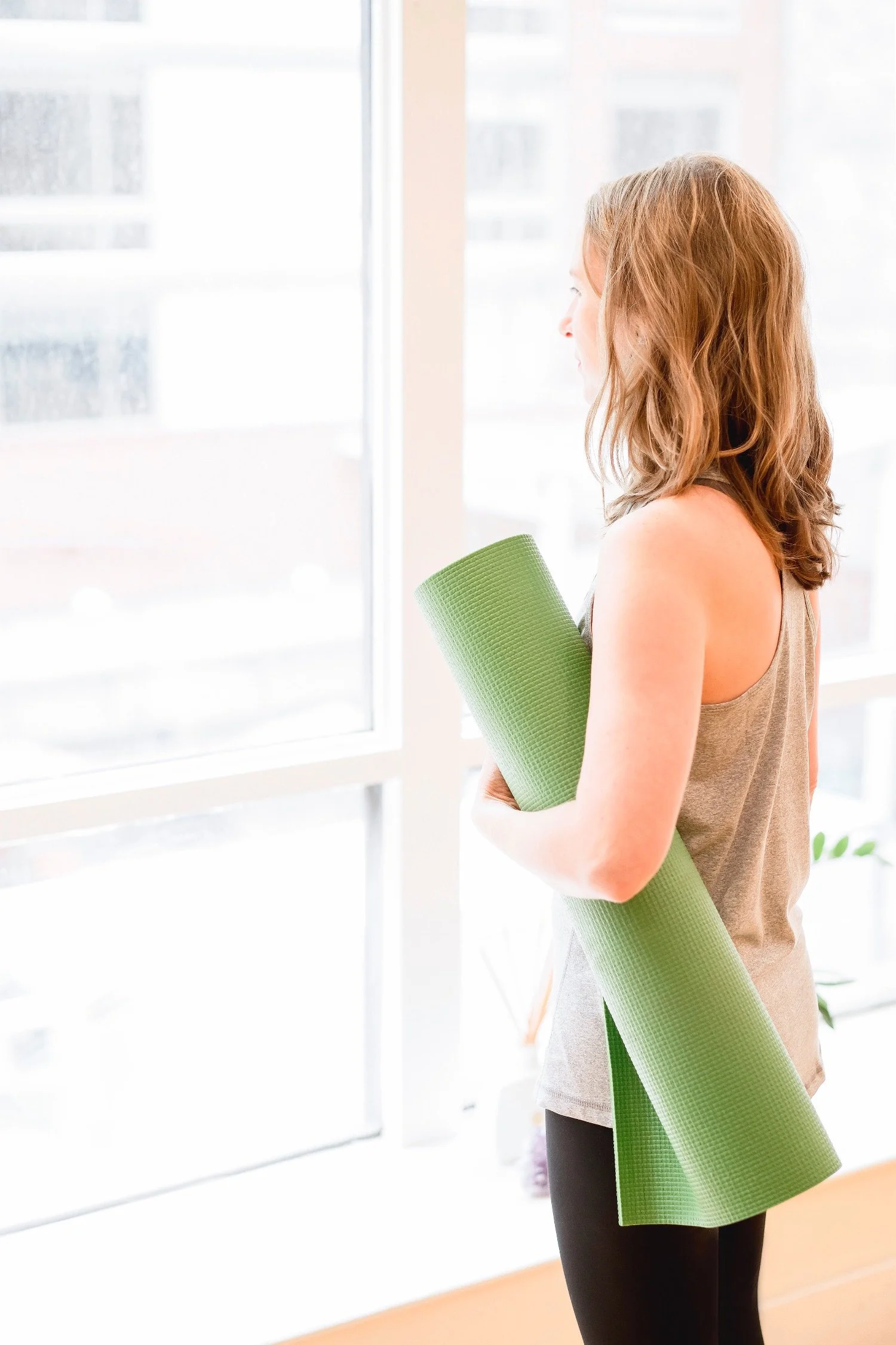 Female wearing gym clothes and carrying a green yoga mat looks out of a large bright window.