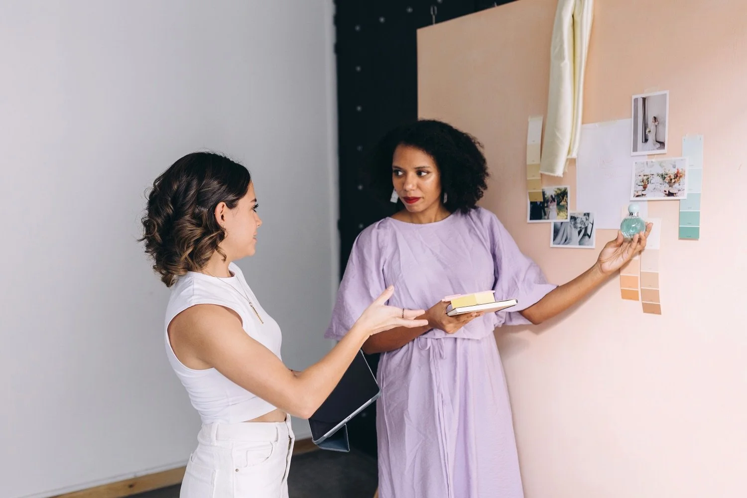 Two females stand at a vision board in a creative office space gesturing towards images on the board.