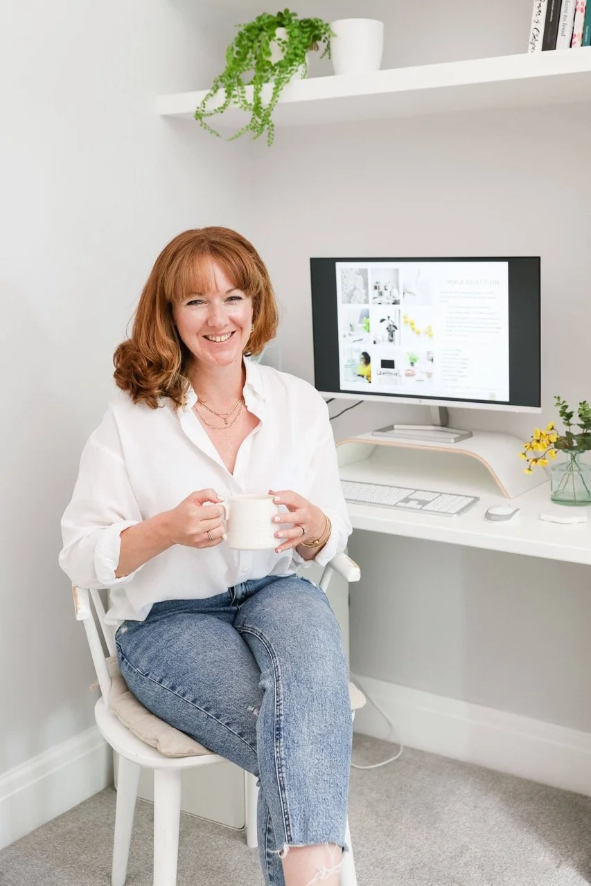 Red haired web designer in jeans and a white shirt sitting holding a mug of tea infront of a desktop monitor and keyboard.