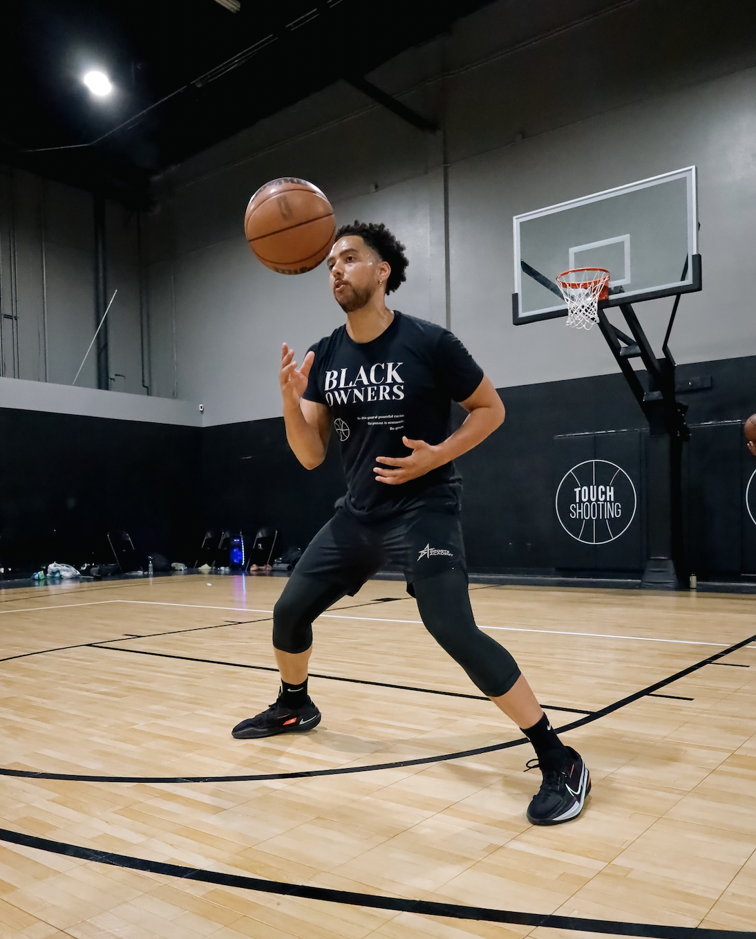 A man in athletic clothing practicing basketball on an indoor court, passing the ball, wearing a black shirt that says 'Black Owners.'