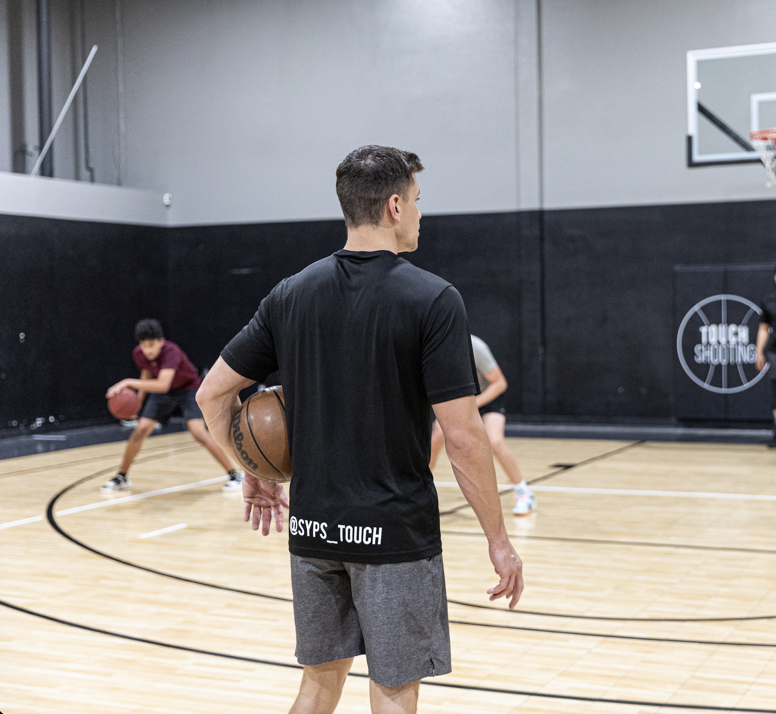 A man coaching children in a basketball gym, holding a basketball, while children practice dribbling in the background.