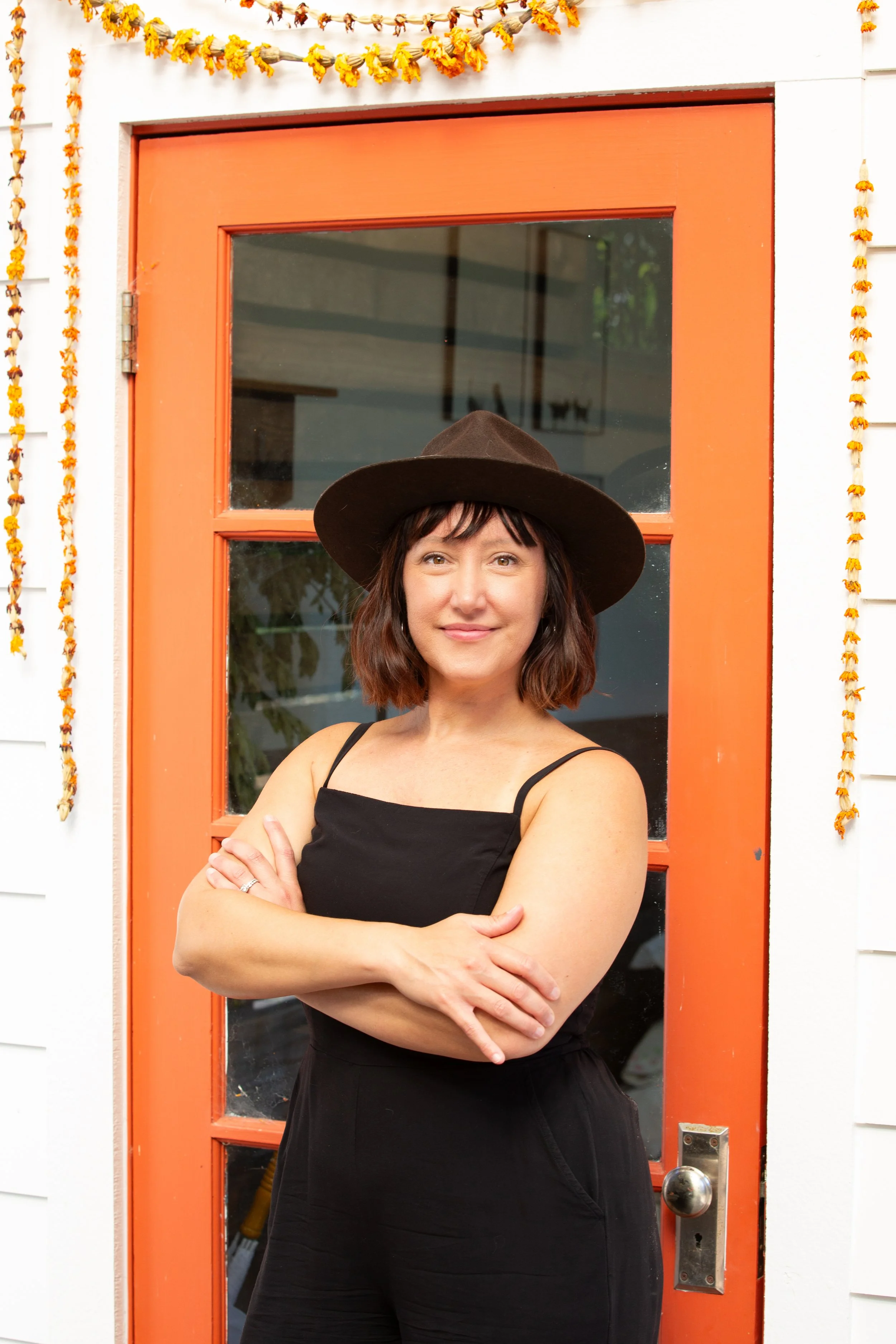 woman wearing black jumper and a hat with arms crossed standing in front of a coral doorway