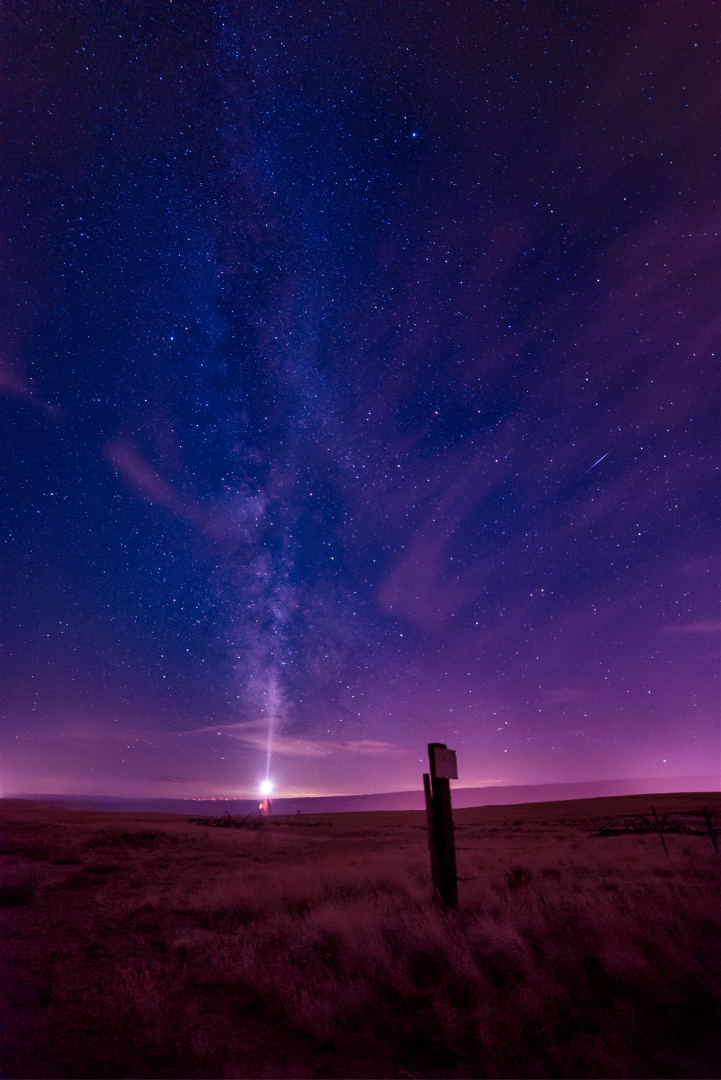 Night sky filled with stars and the Milky Way over an open field, with a bright celestial body emitting a beam of light on the horizon and faint purple clouds.