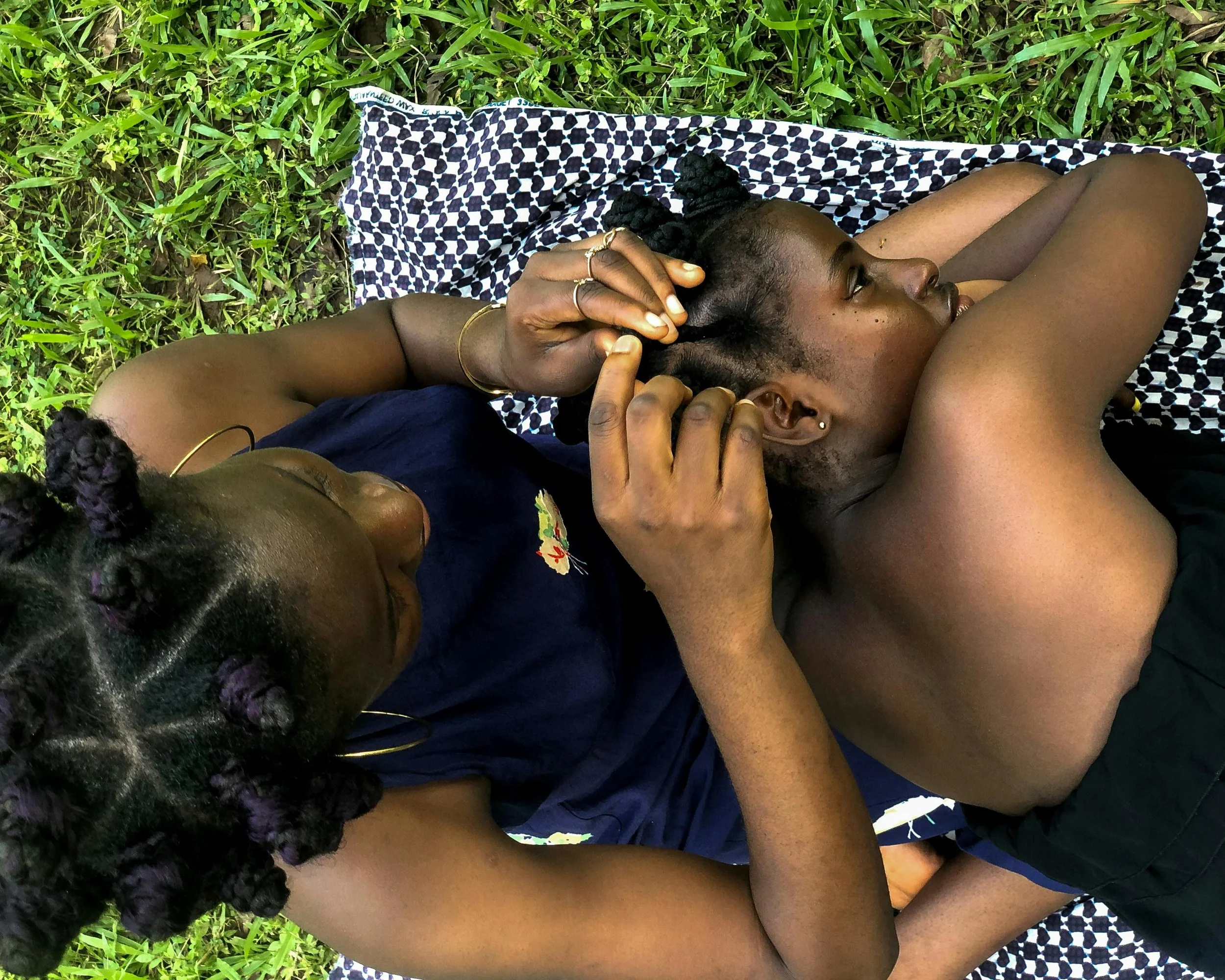 Mulher trança menina em uma fotografia enquadrada de cima, recortando um momento de interação tradicional em povos africanos e afrodescendentes.