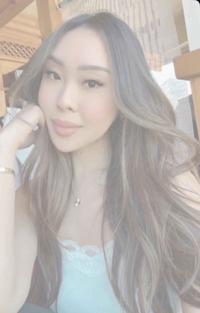 A young woman with long, wavy hair and a light complexion, smiling softly at the camera in an indoor setting with wooden furniture and a bright background.