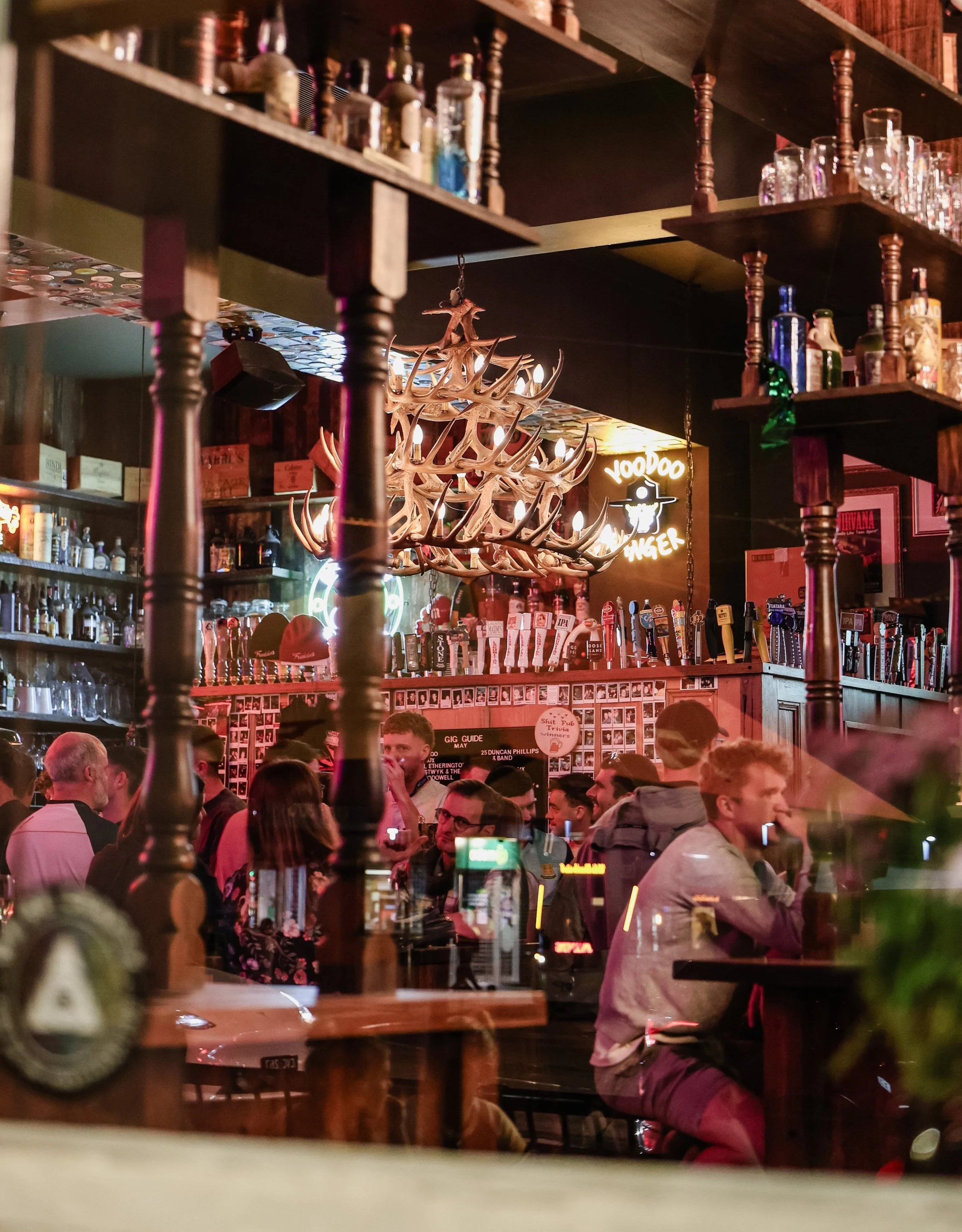 Inside a lively bar filled with people, with an antler chandelier hanging from the ceiling and shelves of liquor bottles. Neon signs and various decor are visible in the background.