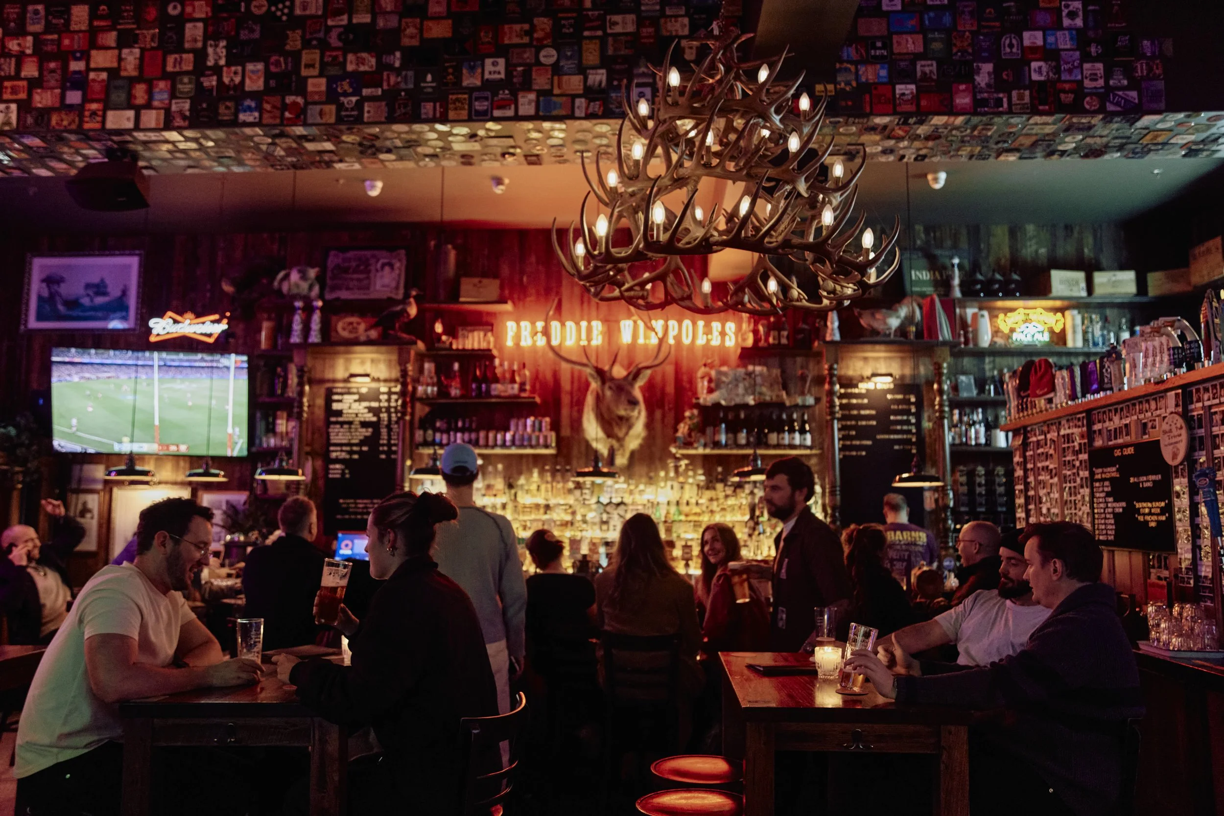 A lively bar scene with people socializing and enjoying drinks. The decor includes a mounted deer head, a large chandelier made of antlers with candle-shaped lights, and a sign that reads 'Freddie Wingo's.' The ceiling is decorated with numerous vintage-style cigarette and matchbook advertisements. A flat-screen TV displays a sports game, and the bar is illuminated with warm lighting.