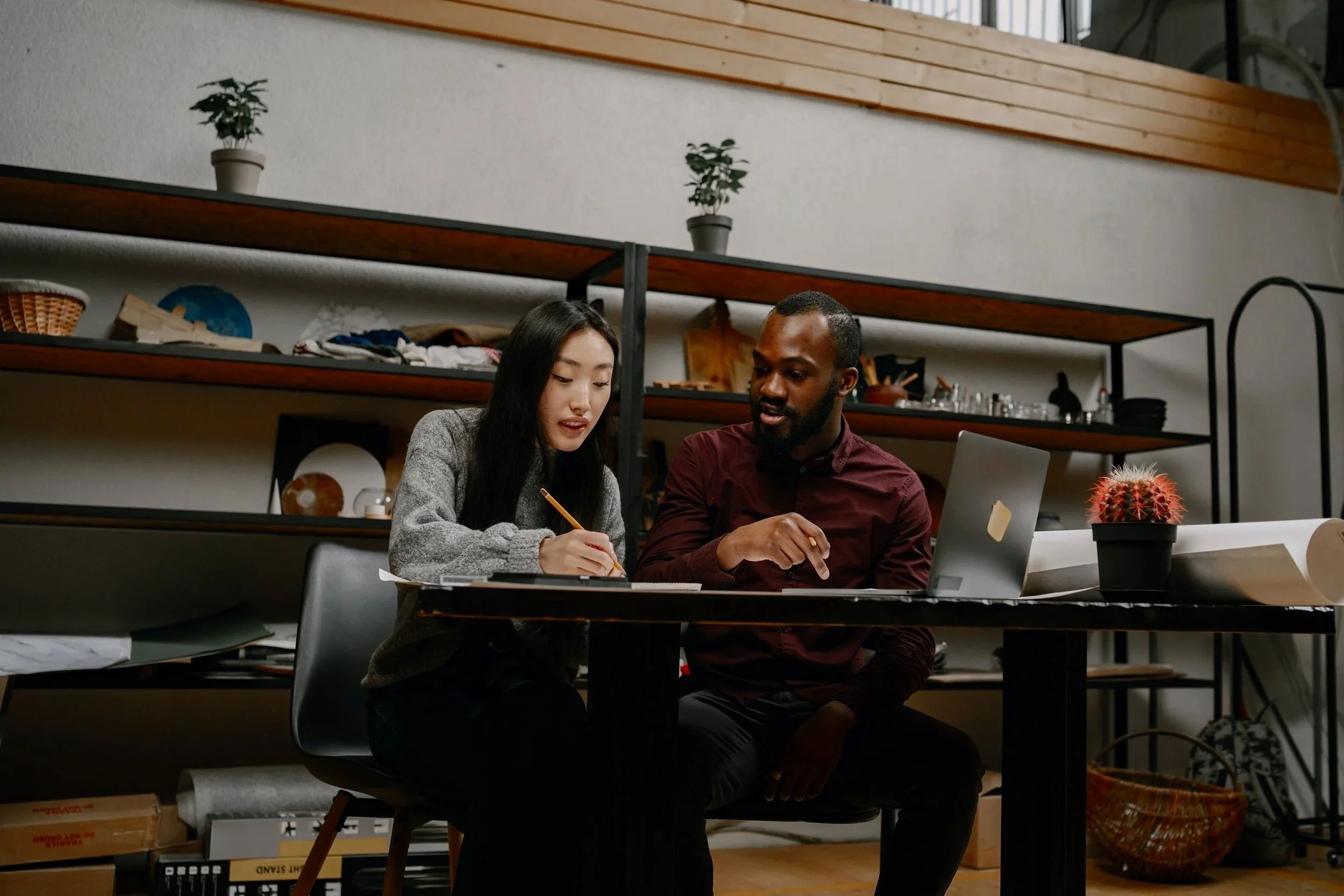 A woman of asian descent and a man of African descent are collaborating on a design. She is drawing and he is using a laptop.