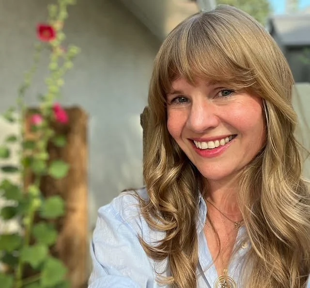 a blond woman with blue eyes smiles at the camera, She has long hair with bangs and is wearing a blue collared shirt.
