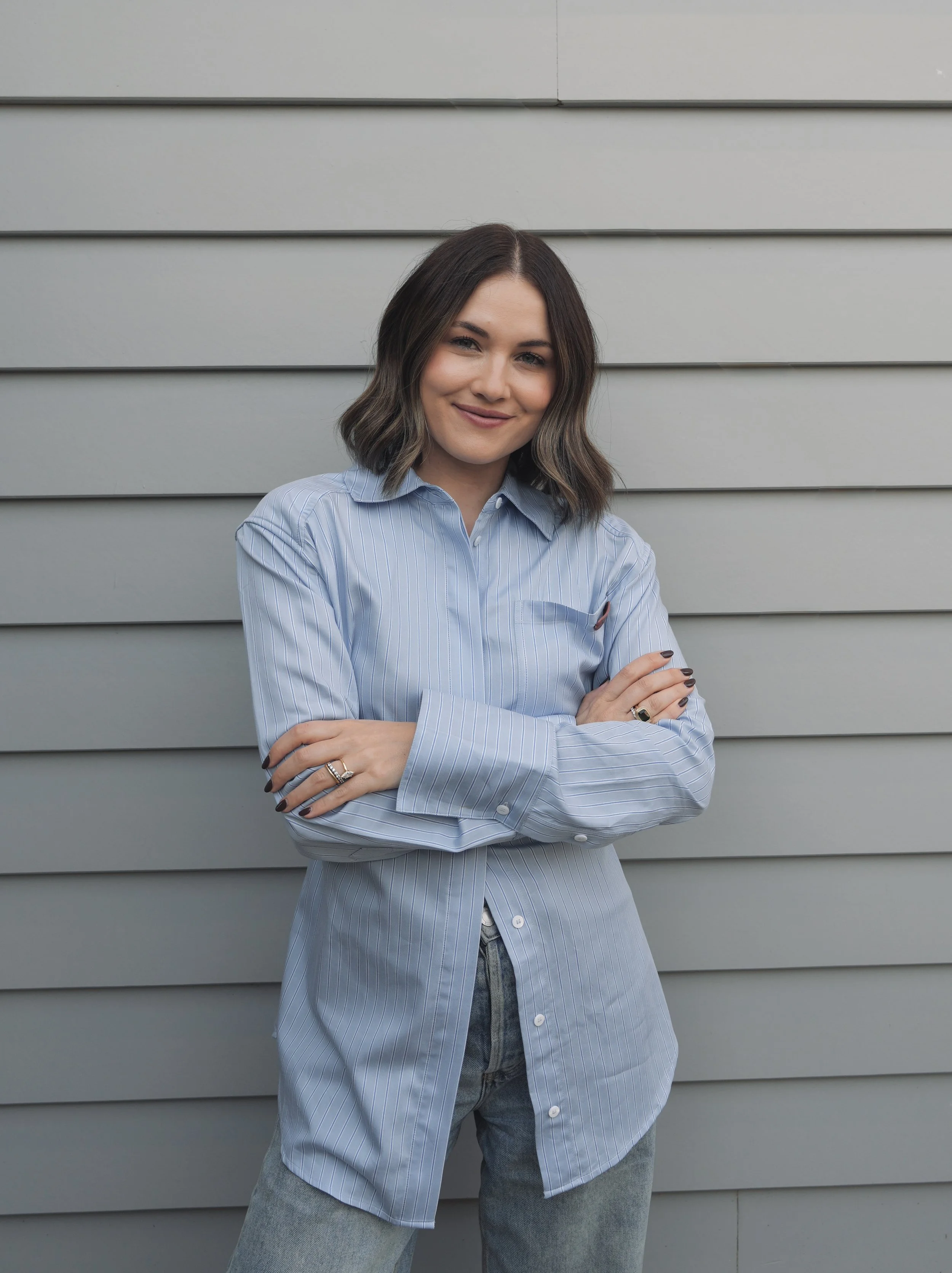 A woman standing with arms crossed, smiling, wearing a light blue striped button-up shirt and gray jeans, standing against a gray horizontal siding wall.