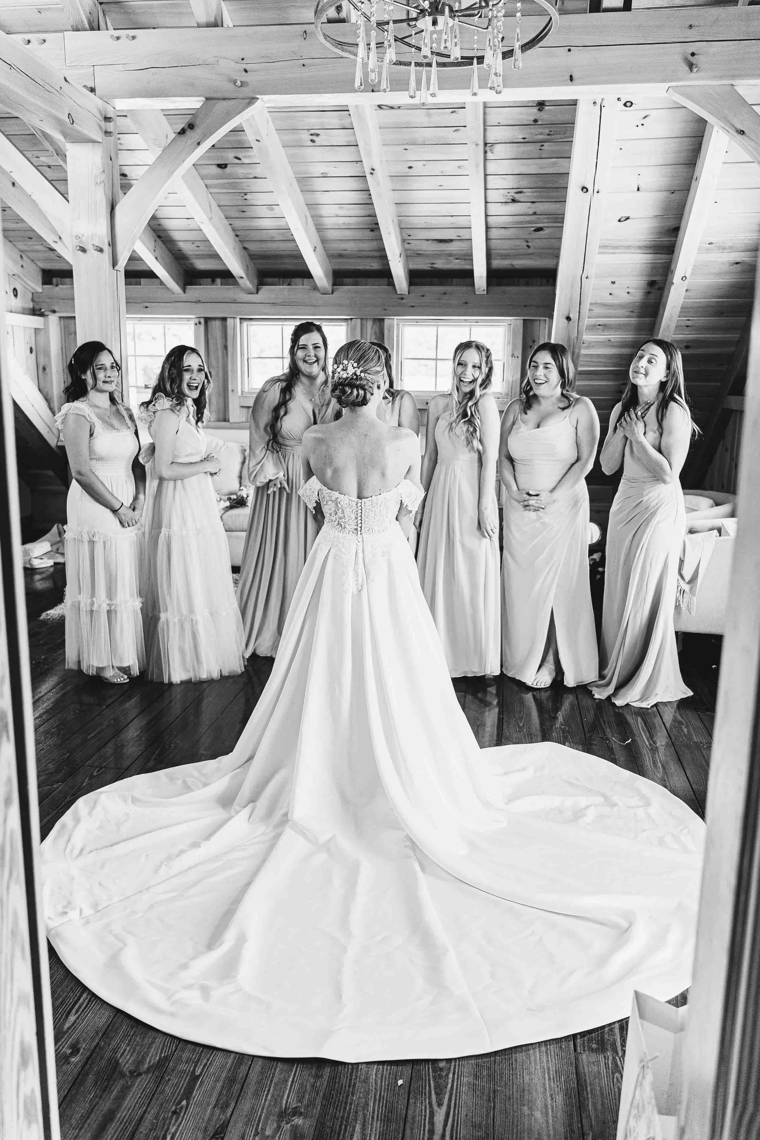 A bride in a wedding dress and veil faces her bridesmaids, who are wearing light-colored dresses, inside a rustic wooden room with a chandelier.