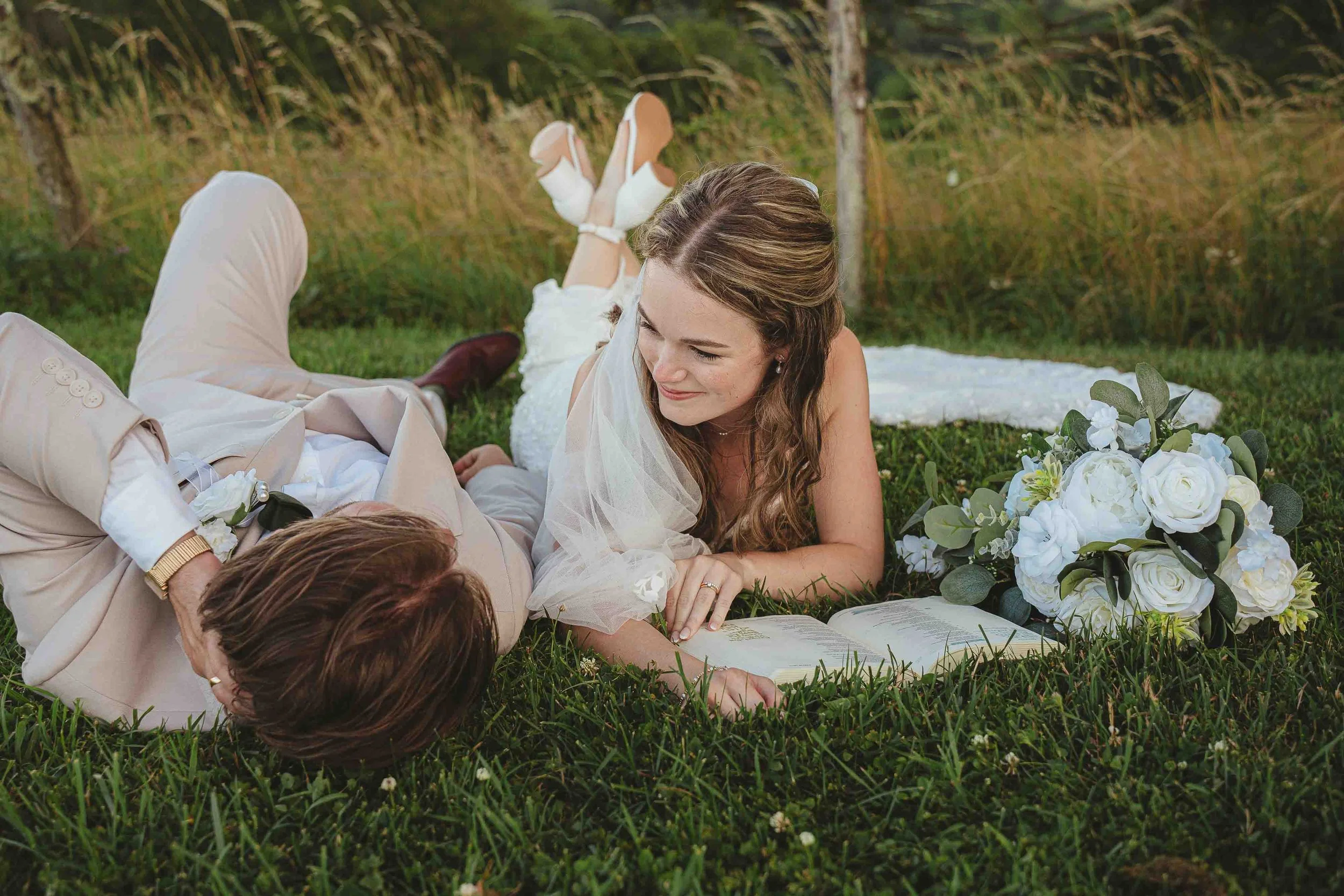 A woman and a man lying on grass outdoors with a white bouquet and an open book, possibly a wedding or engagement scene.