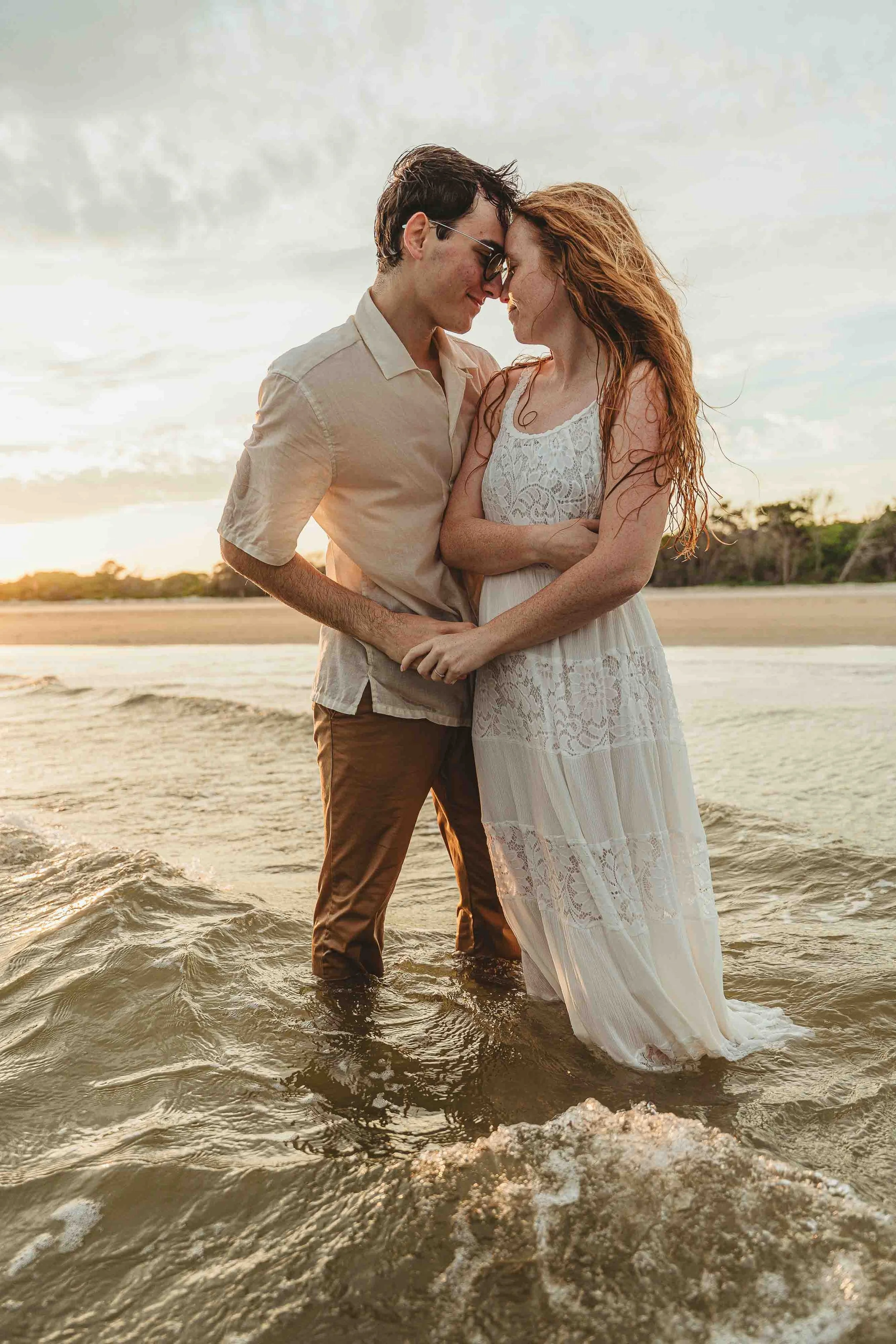 A couple standing together in shallow water at the beach during sunset, gazing into each other's eyes and smiling, with the woman wearing a white lace dress and the man wearing a light-colored shirt and brown pants.