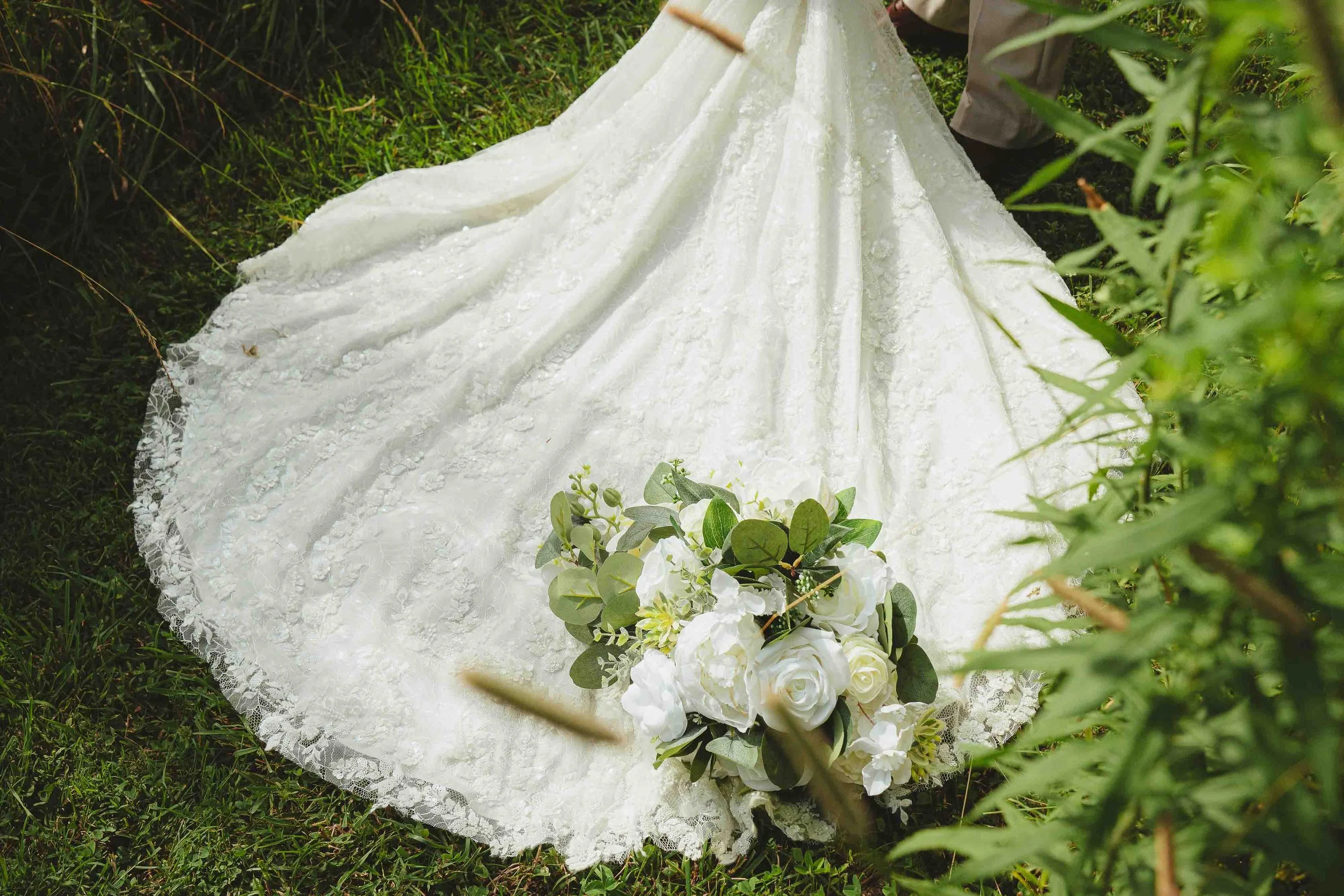 A white lace wedding dress on the grass with a bouquet of white roses and greenery.