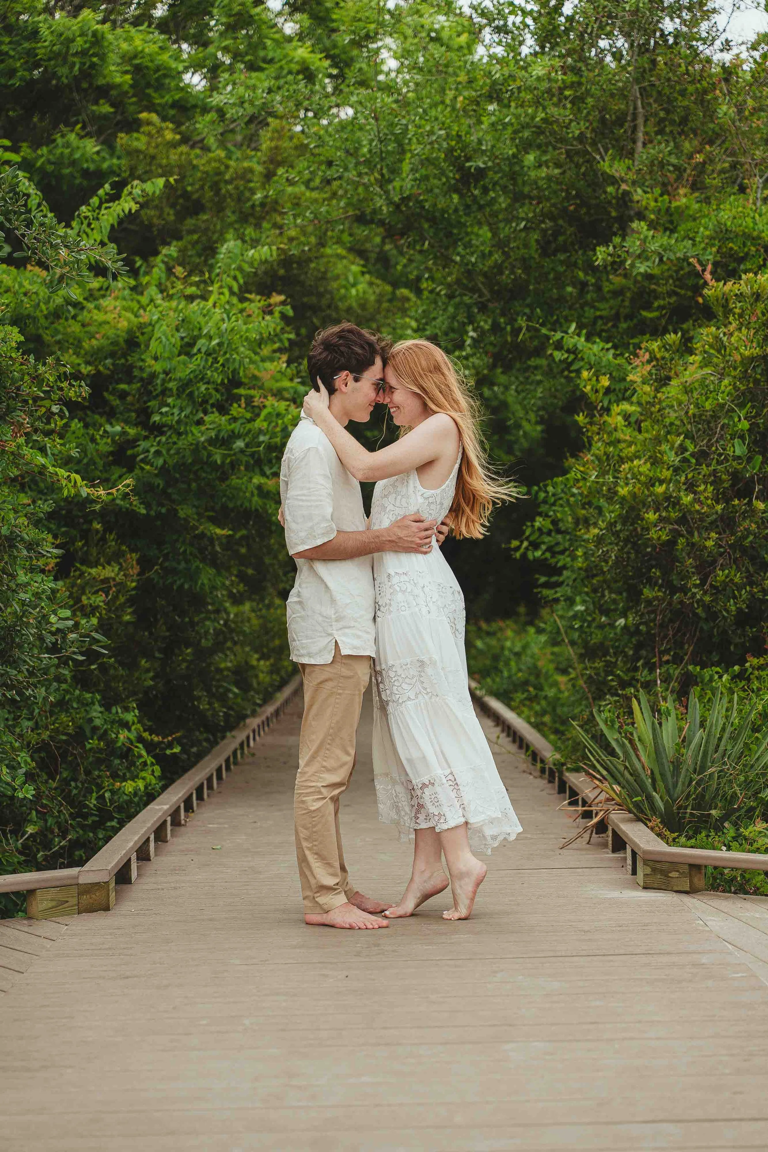 A couple stands close with foreheads touching and eyes closed on a wooden bridge surrounded by lush green foliage, both barefoot, dressed in light-colored clothing, sharing an intimate moment.
