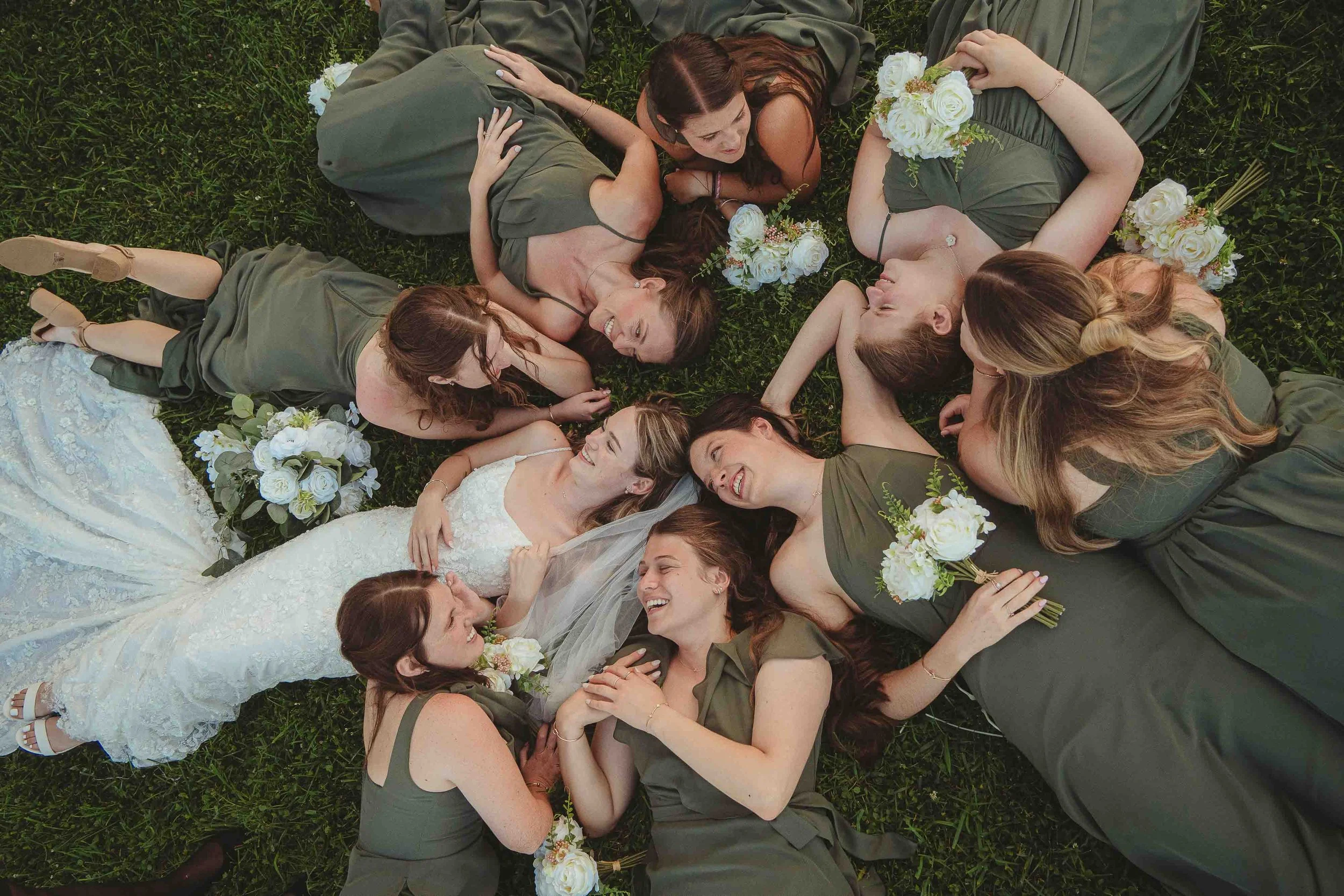 A group of women, including a bride in a white dress, lying on the grass in a circle, smiling and looking at each other, surrounded by bouquets of white and light pink flowers.