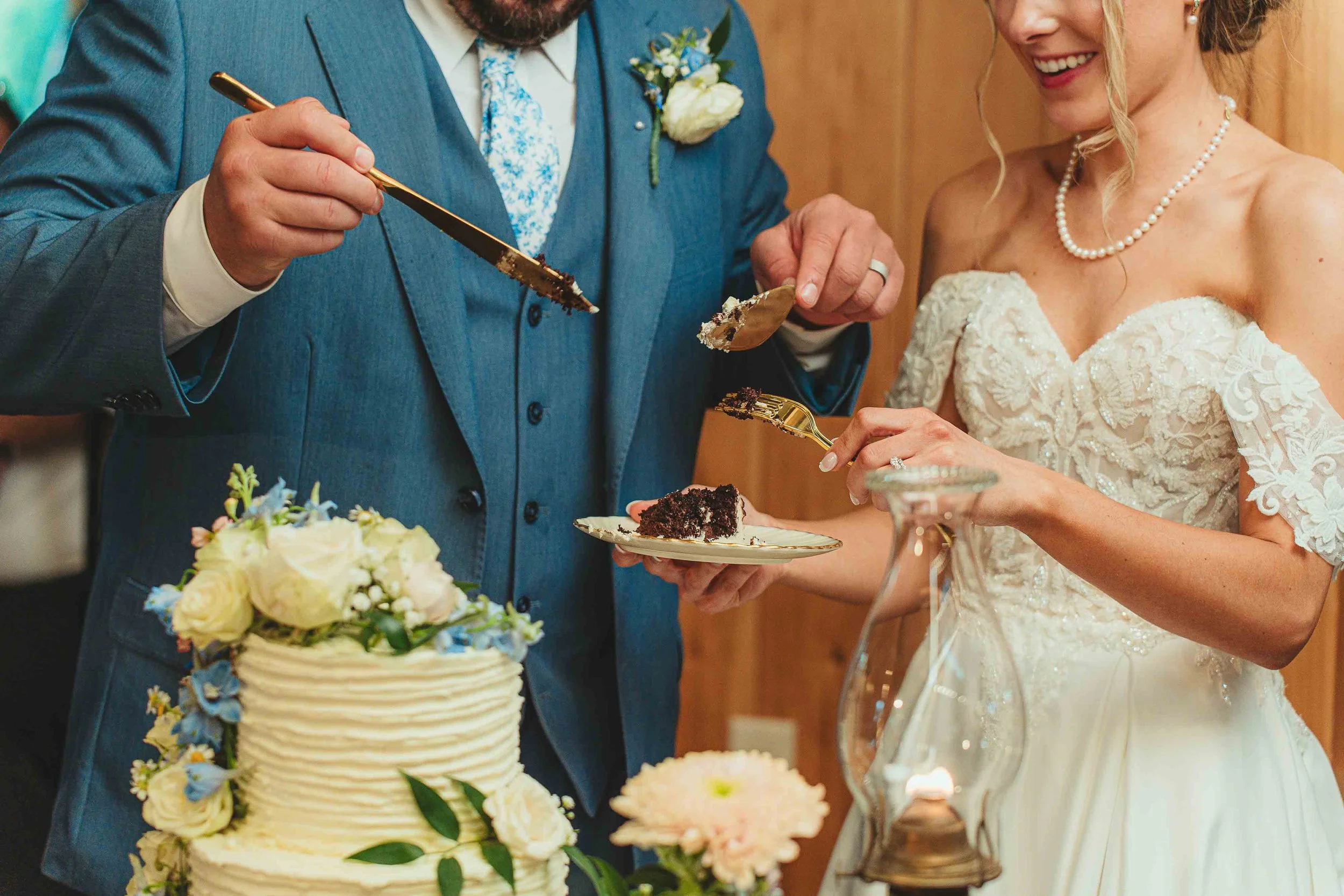 A bride and groom enjoying a slice of wedding cake during their wedding celebration, with a decorated cake in front of them and floral arrangements nearby.