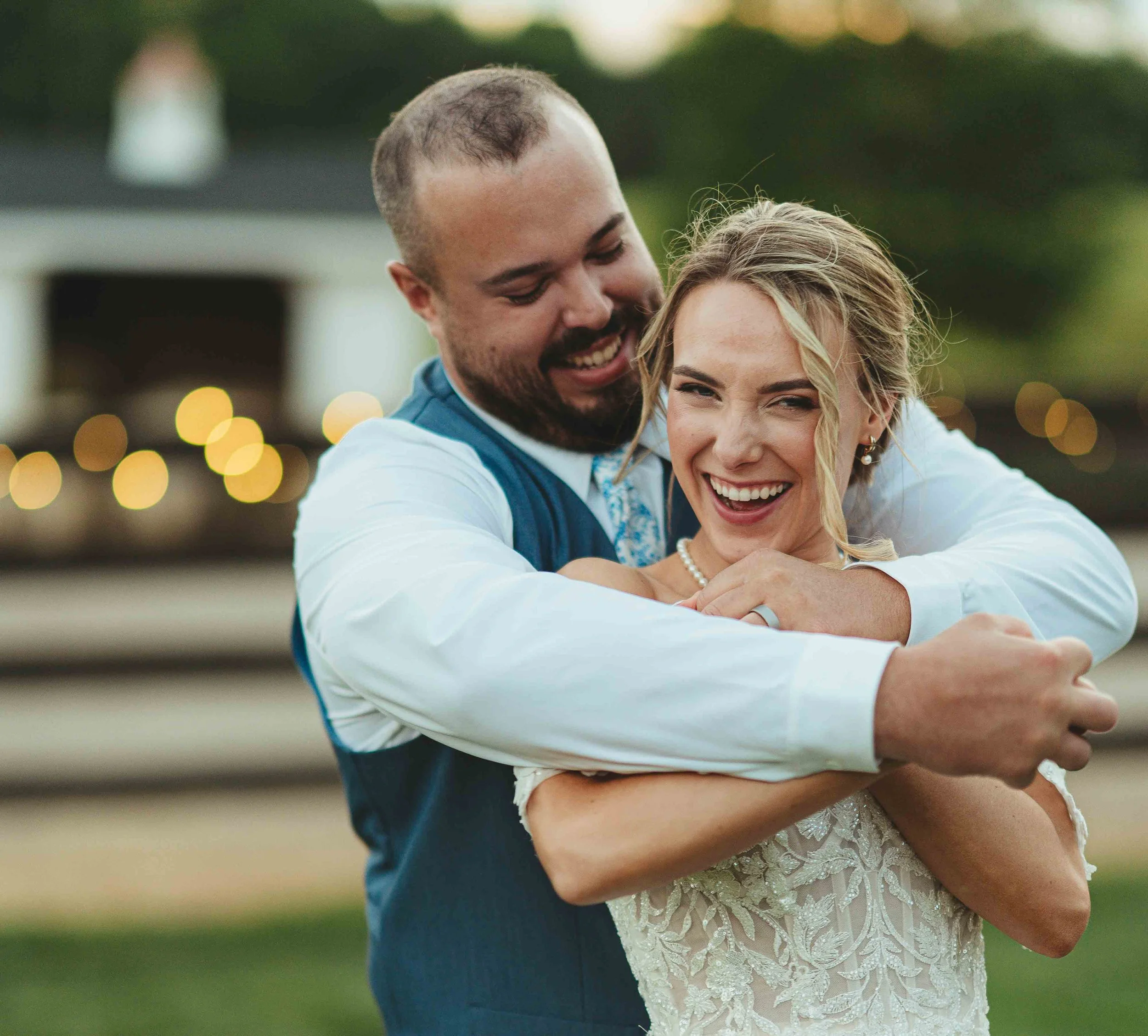 A happy couple, dressed in wedding attire, sharing an embrace outdoors during sunset, with bokeh lights in the background.