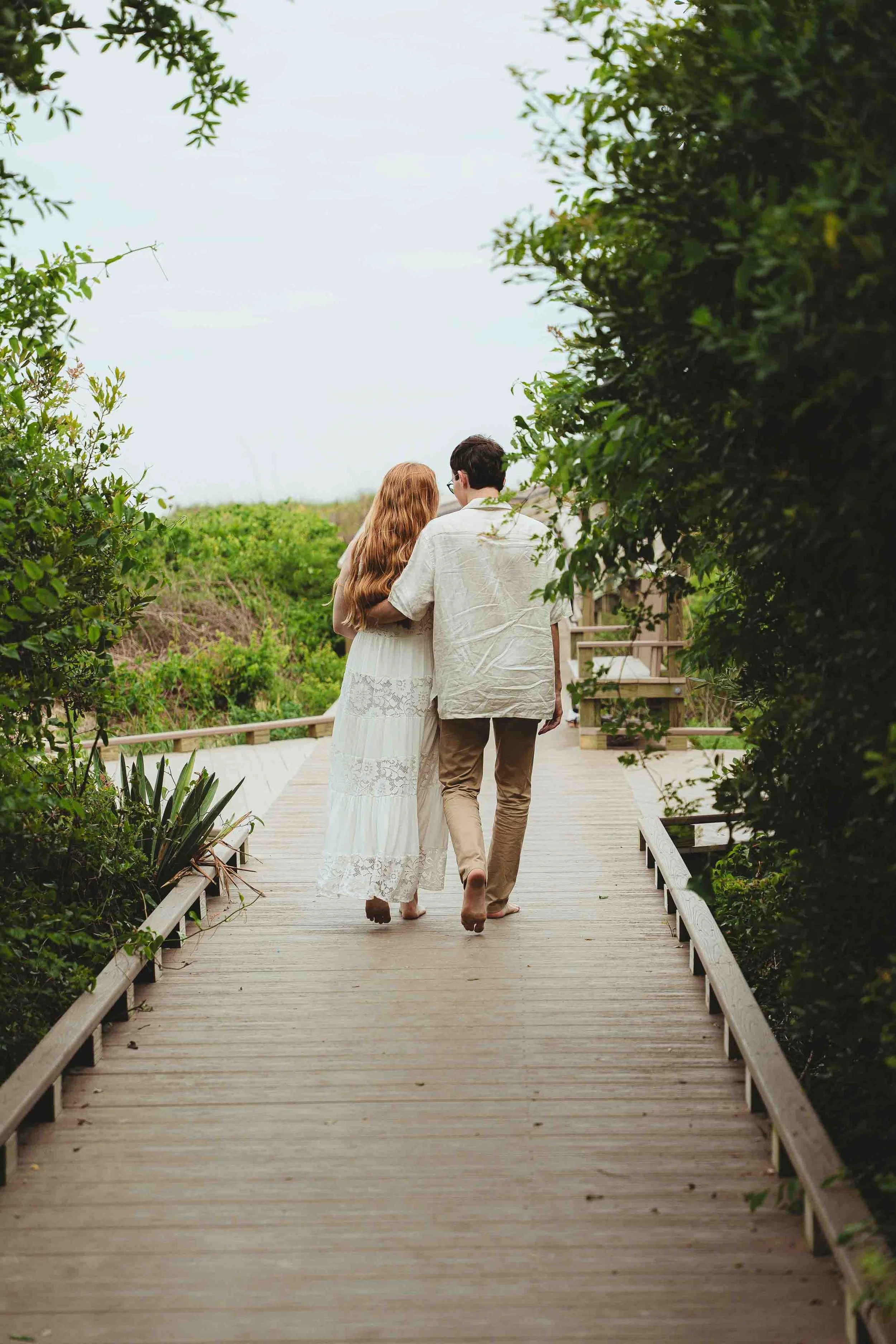 A couple walking on a wooden pathway surrounded by greenery, with the woman having long red hair and the man with dark hair, both dressed in light-colored clothing.