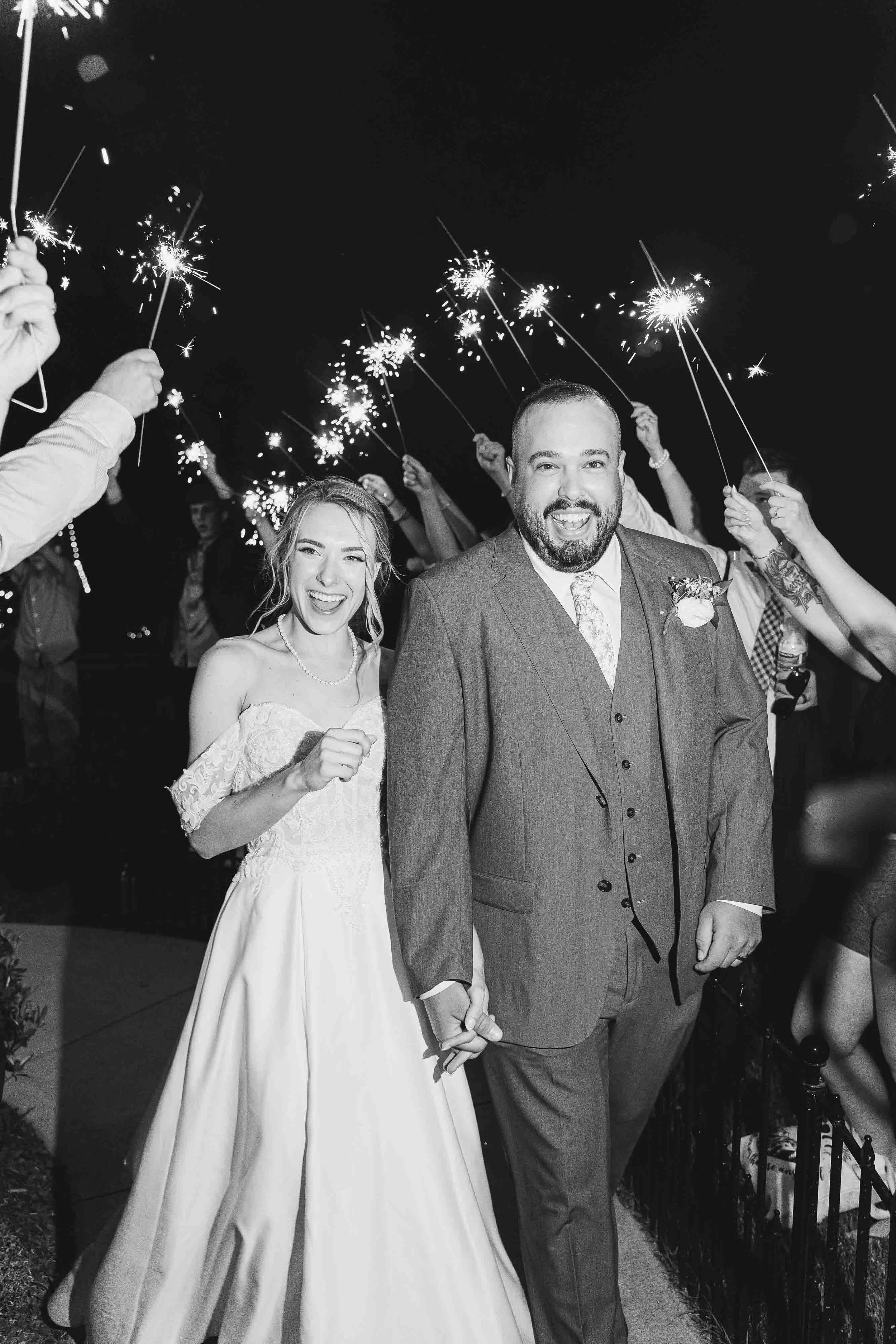 A newlywed couple holding hands and smiling at night, surrounded by friends holding sparklers in celebration.