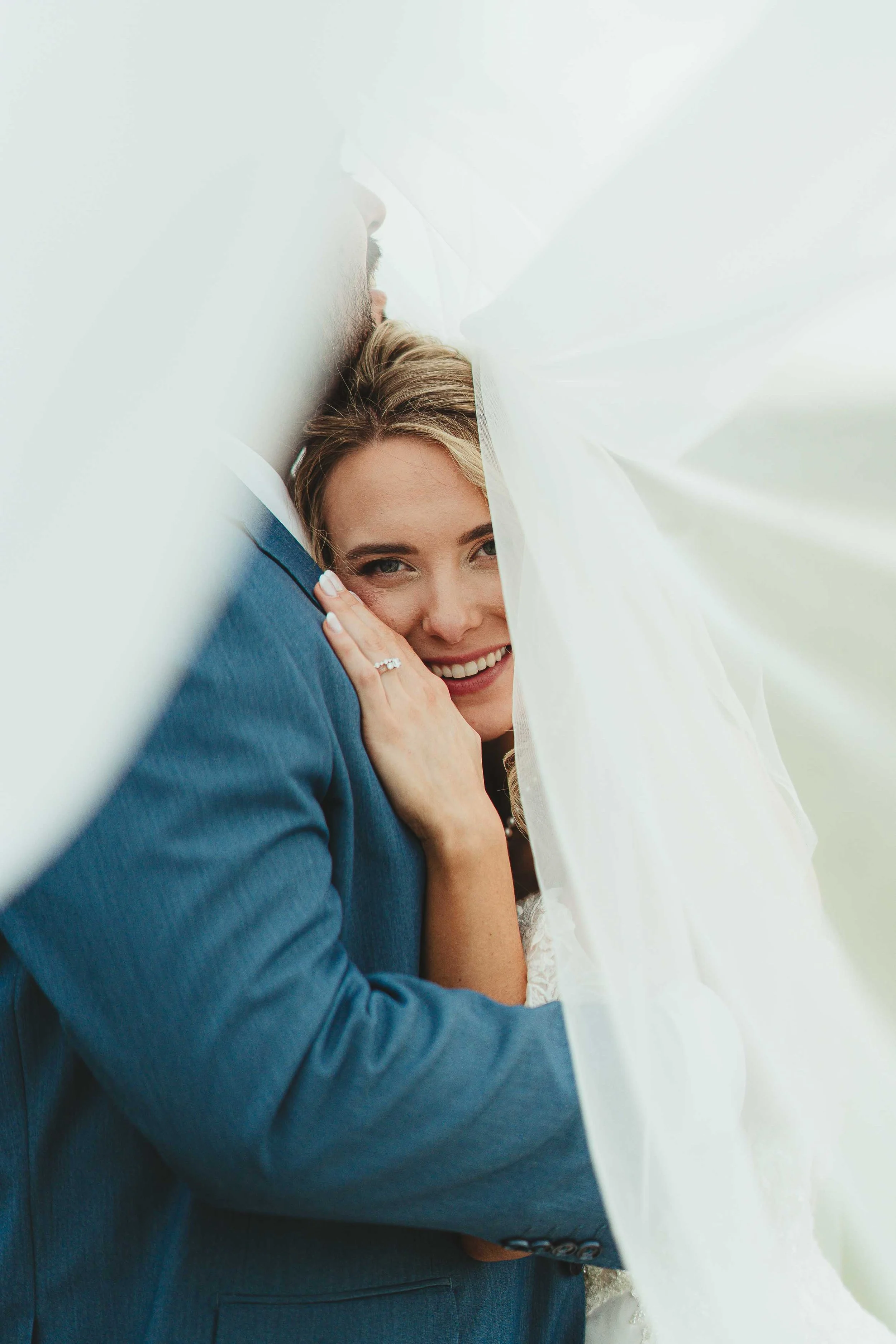 Close-up of a smiling woman with blonde hair, hugging a man in a blue suit, partially covered by white fabric.