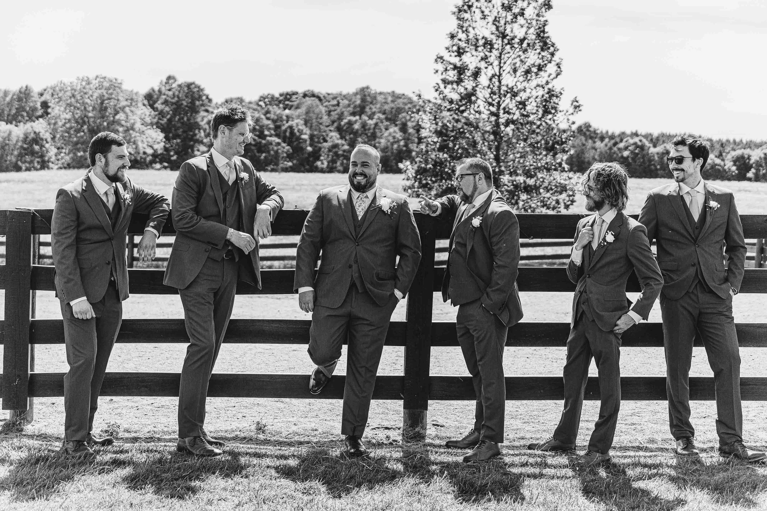 A group of seven men in suits standing outside near a wooden fence, engaging in conversation and smiling, with a background of trees and open sky.