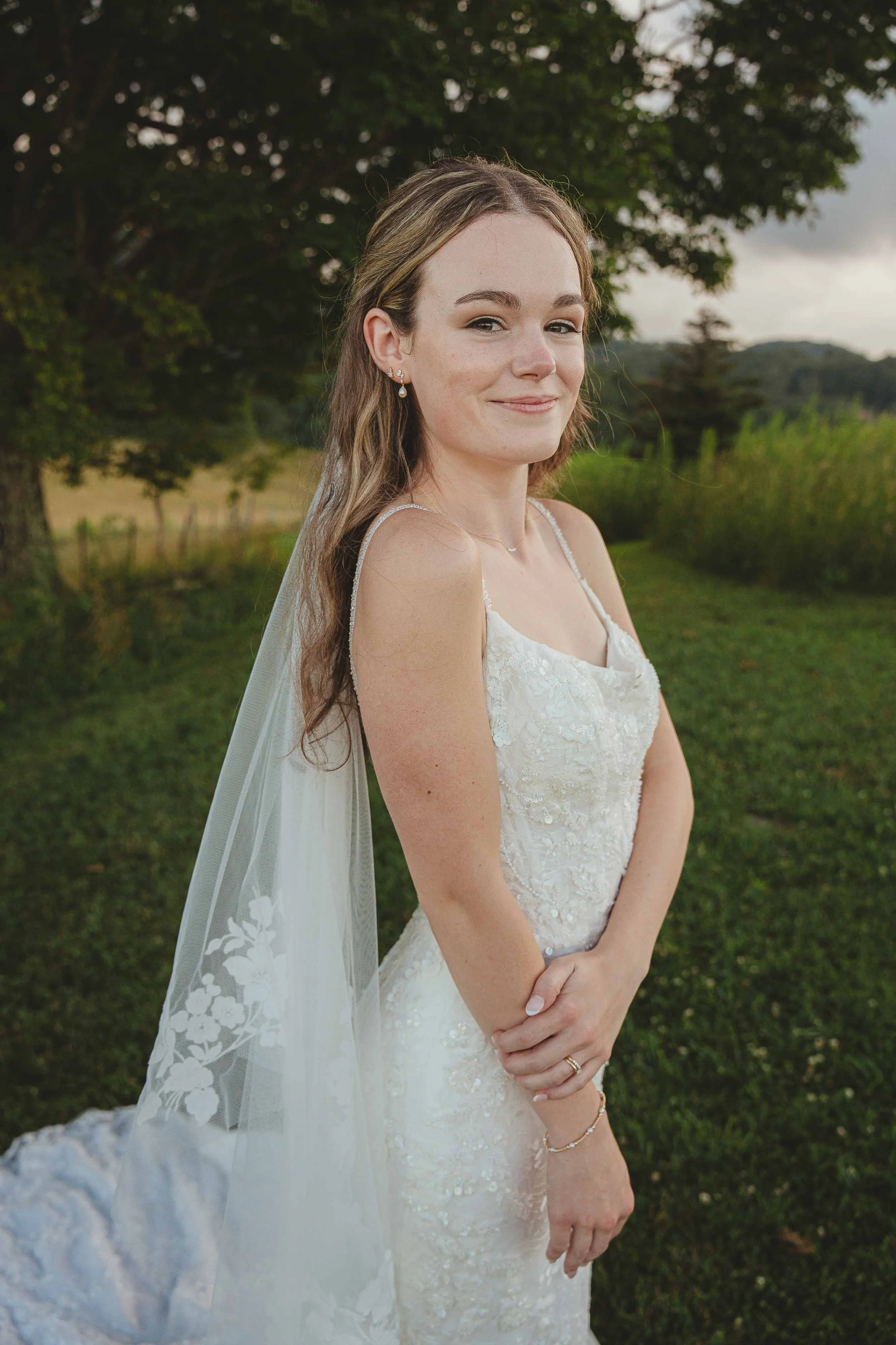 A bride in a white wedding dress with lace details and spaghetti straps, standing outdoors on a grassy field near trees, with a scenic background and cloudy sky.