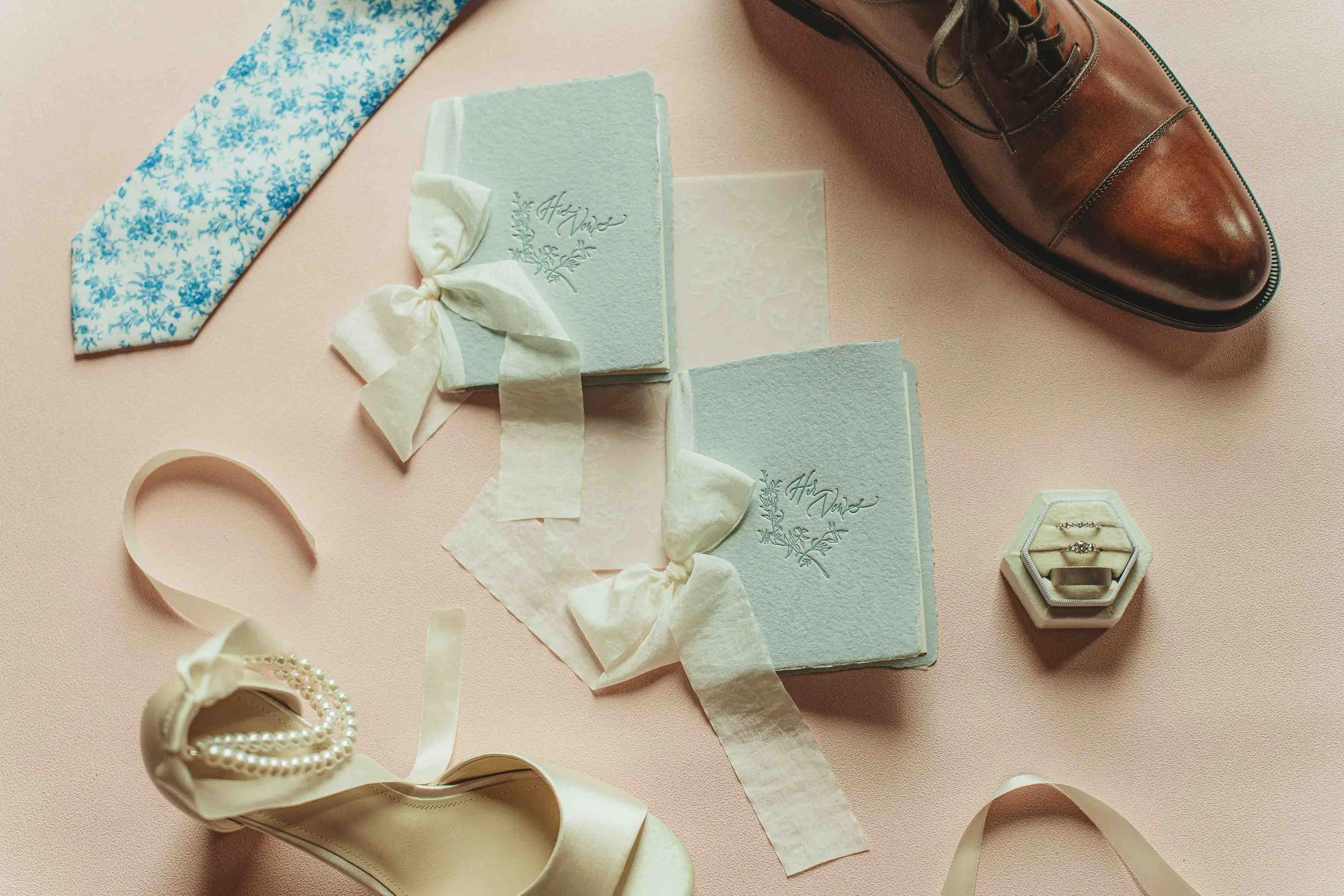 Wedding accessories on a pink surface, including a brown leather dress shoe, a pair of wedding rings in a hexagonal ring box, a white satin shoe with pearls, a light blue wedding guest book with ribbon, a blue floral tie, and a small piece of delicat