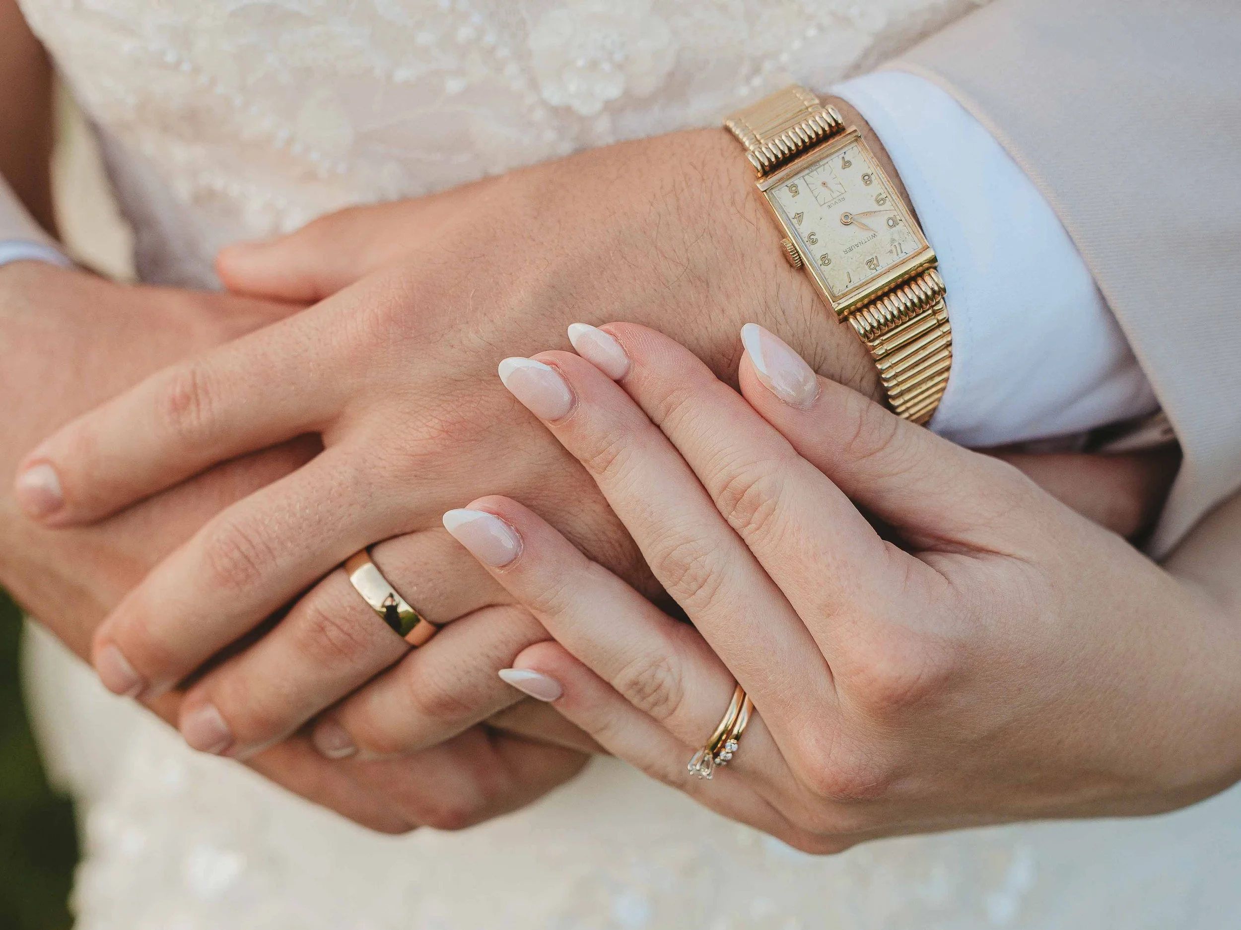 Close-up of a woman and man holding hands, showing wedding rings. The man wears a gold wristwatch and a wedding band, while the woman has a diamond ring on her finger. They are dressed in formal attire, likely for a wedding.