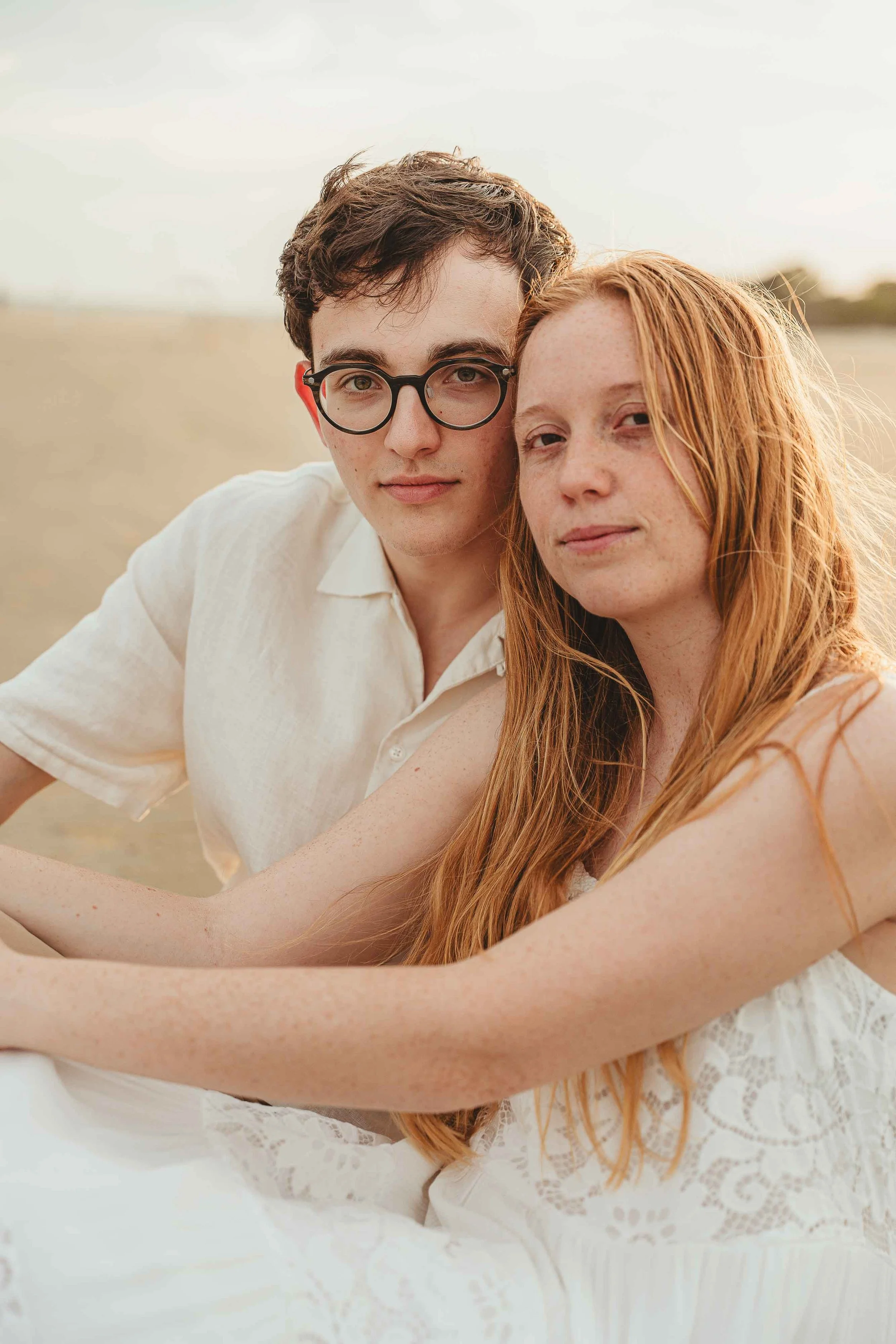 A young man with glasses and a young woman with long red hair sitting close together outdoors during sunset.