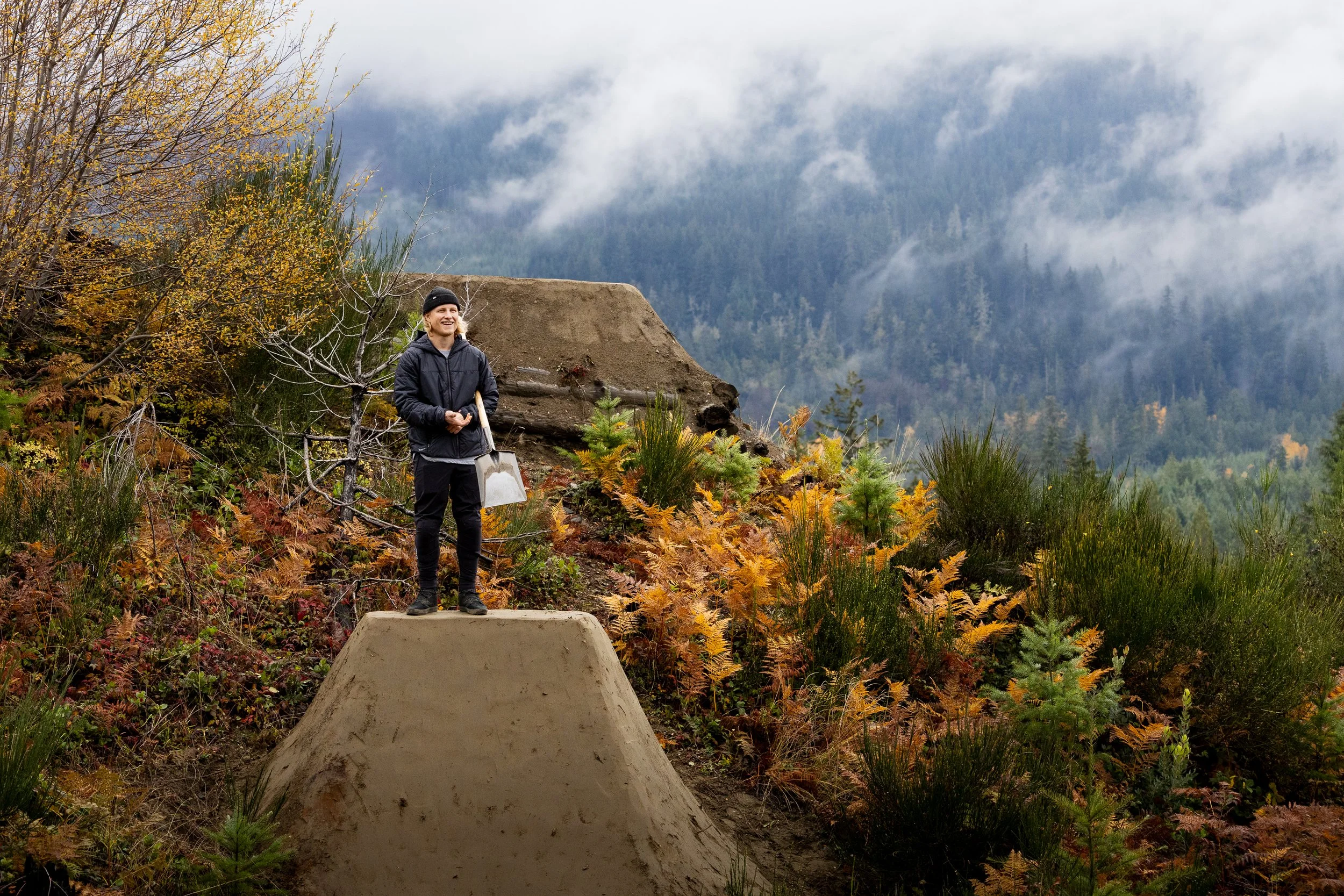 A person standing on a dirt ramp in a forested mountainous area, holding a shovel and smiling, with autumn foliage and mountain peaks in the background.