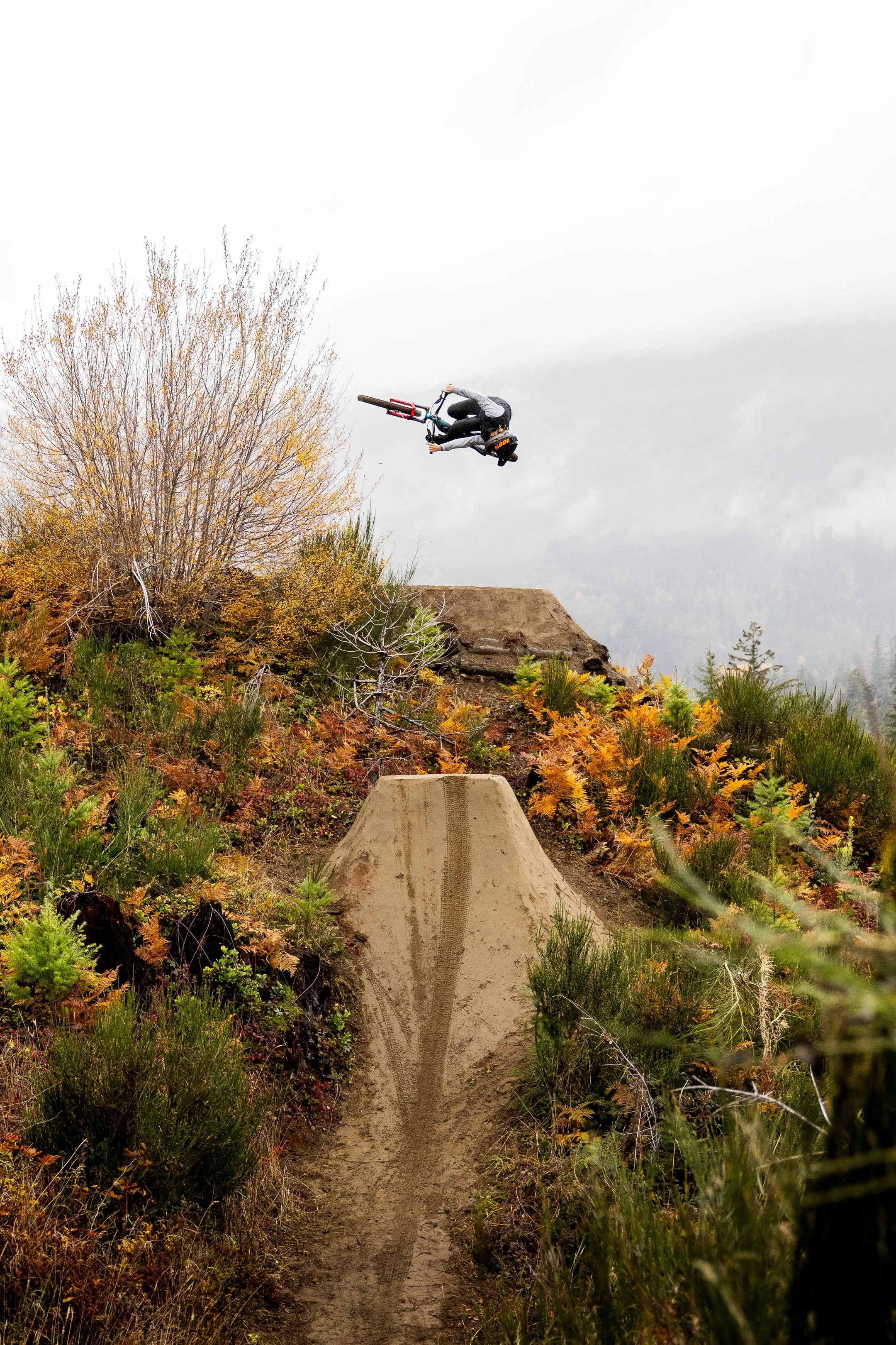 Mountain biker mid-air executing a trick on a dirt ramp in a forested area with autumn foliage.