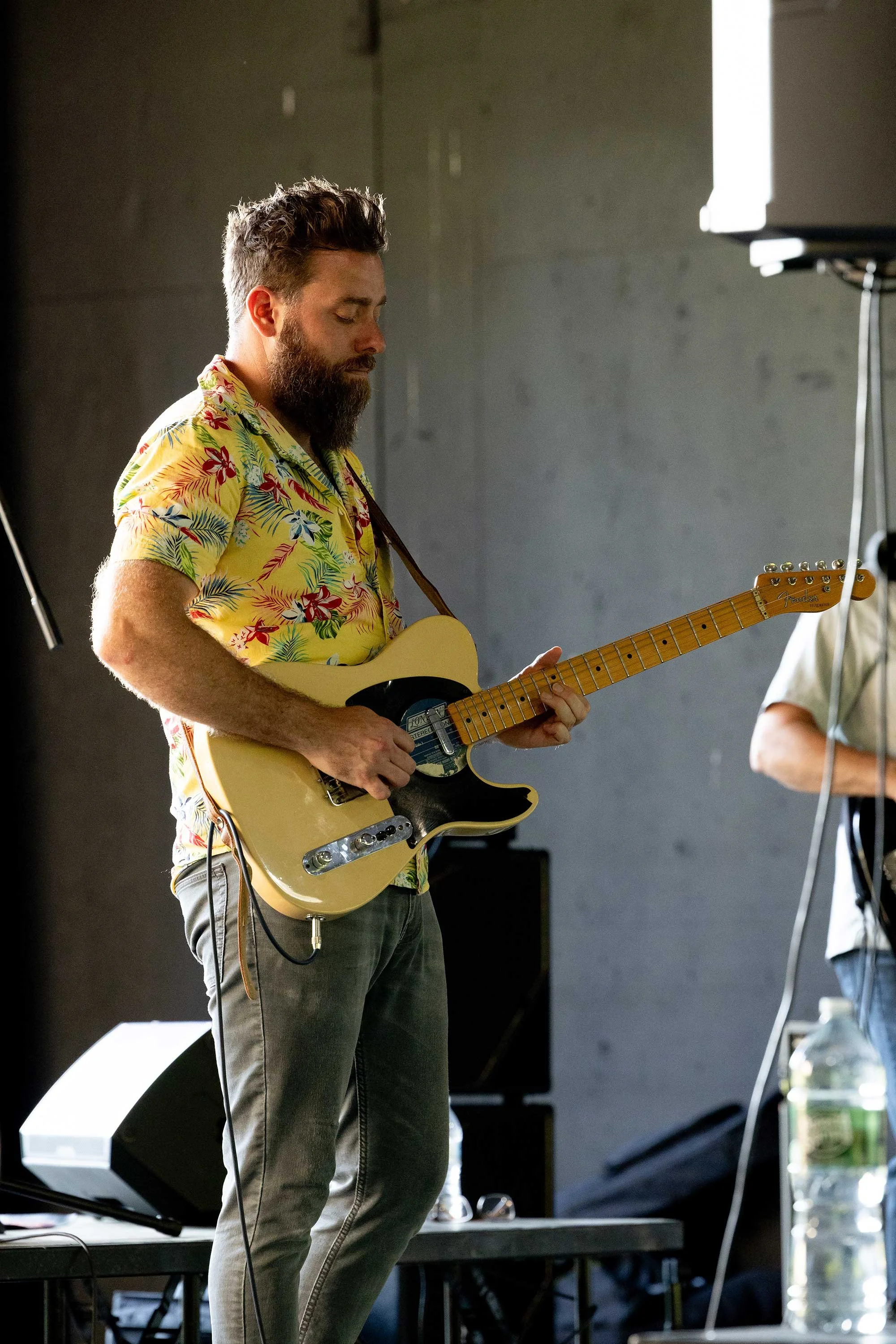 A man with a beard and dark hair is playing an electric guitar. He is wearing a yellow Hawaiian shirt with floral patterns and light gray jeans. He stands on a stage with a dark background and some equipment nearby.