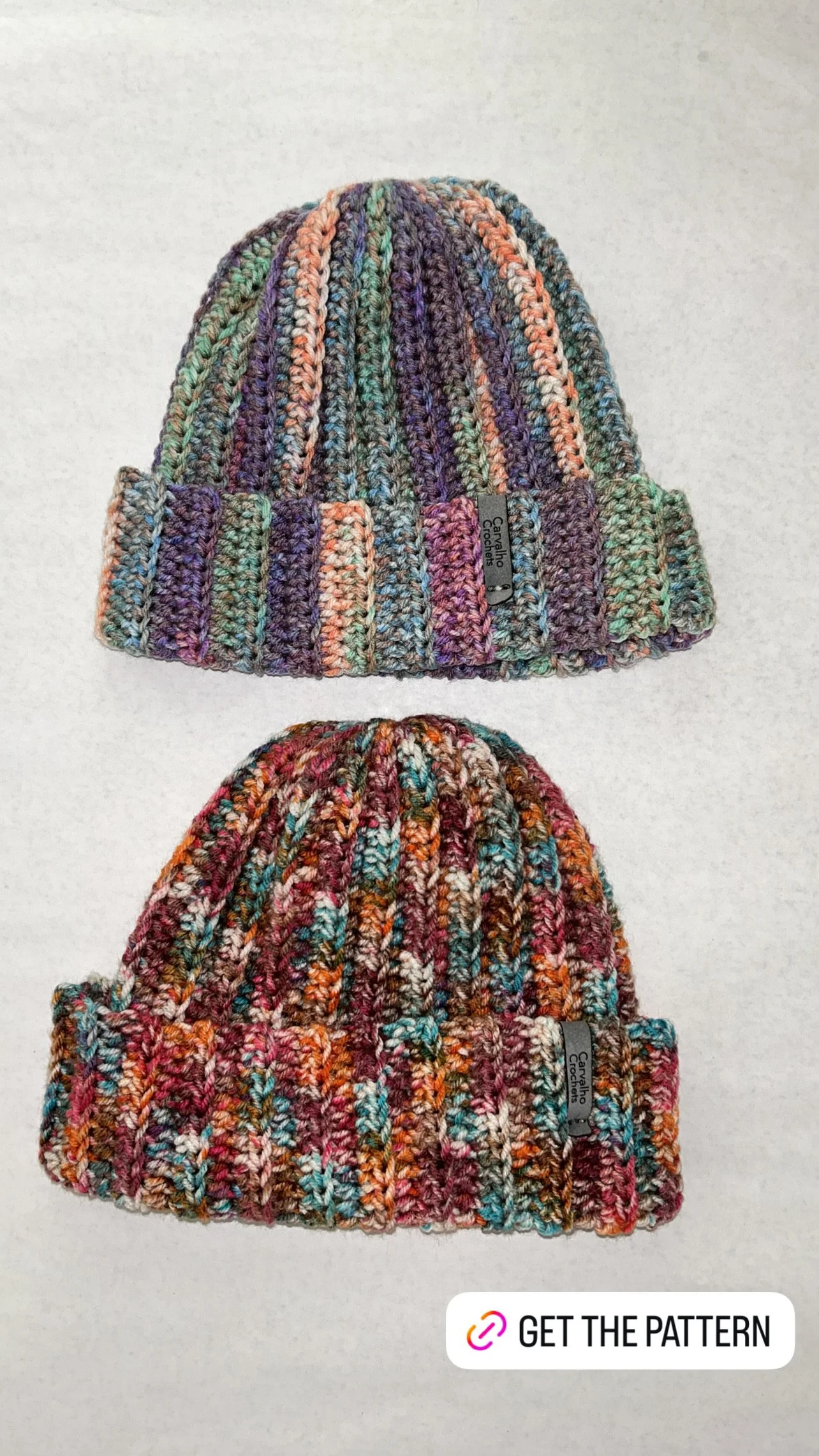 Two colorful knit beanies, one with purple, green, and peach tones, and the other with red, orange, and blue tones, placed one above the other on a white background.