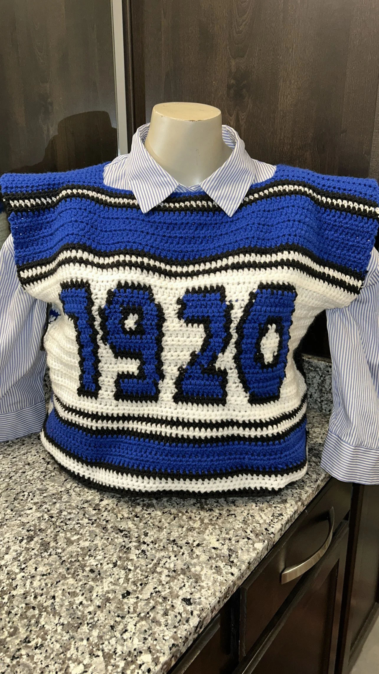 A crocheted vintage-style sports jersey with the number 1920, layered over a striped dress shirt, positioned on a kitchen countertop.