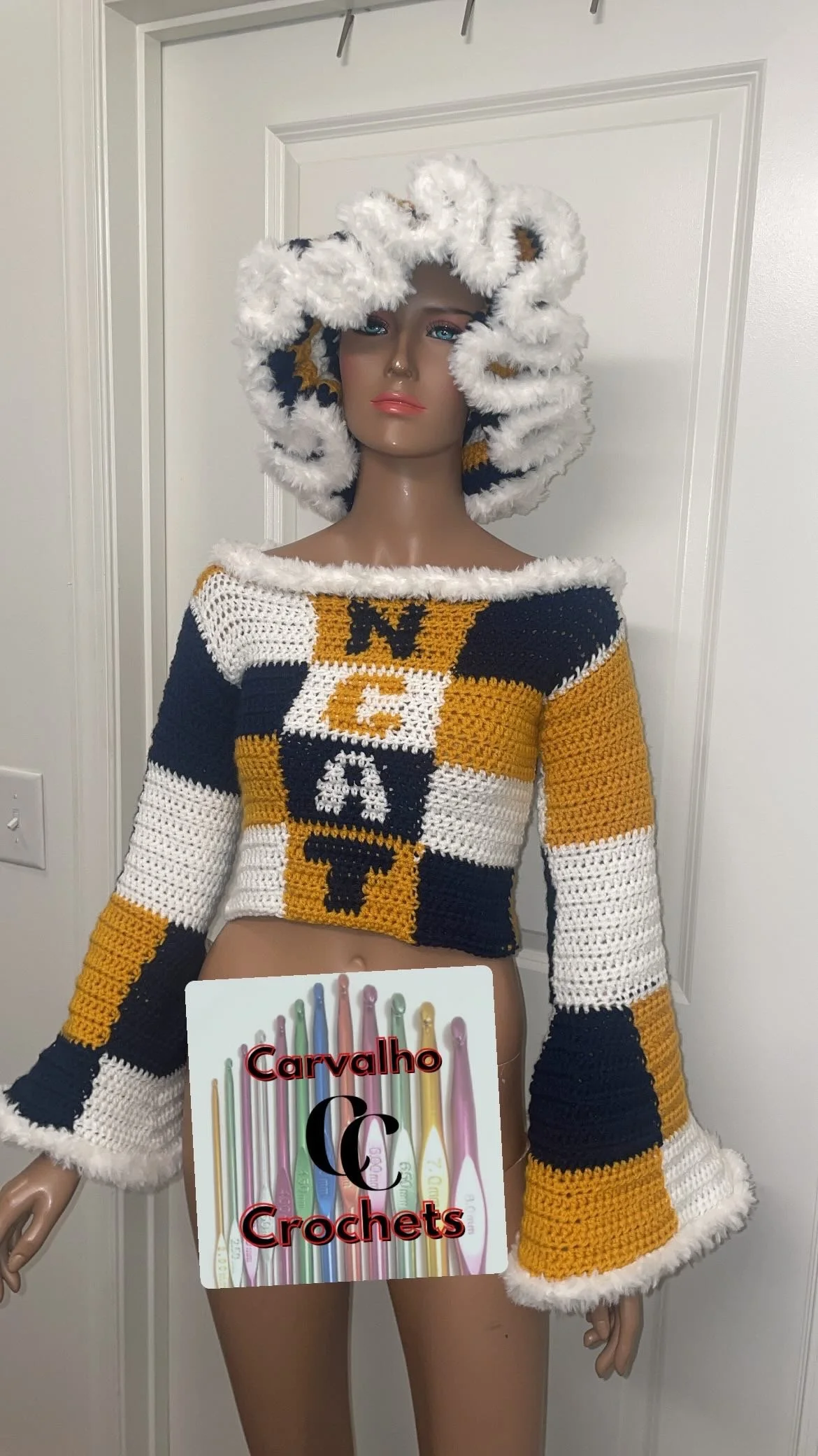 A mannequin dressed in a crocheted sweater with a checkered pattern of navy blue, white, and mustard yellow, and a matching hood. The mannequin is standing in front of a white door. There is a sign on the mannequin's waist reading 'Carvalho Crochets'