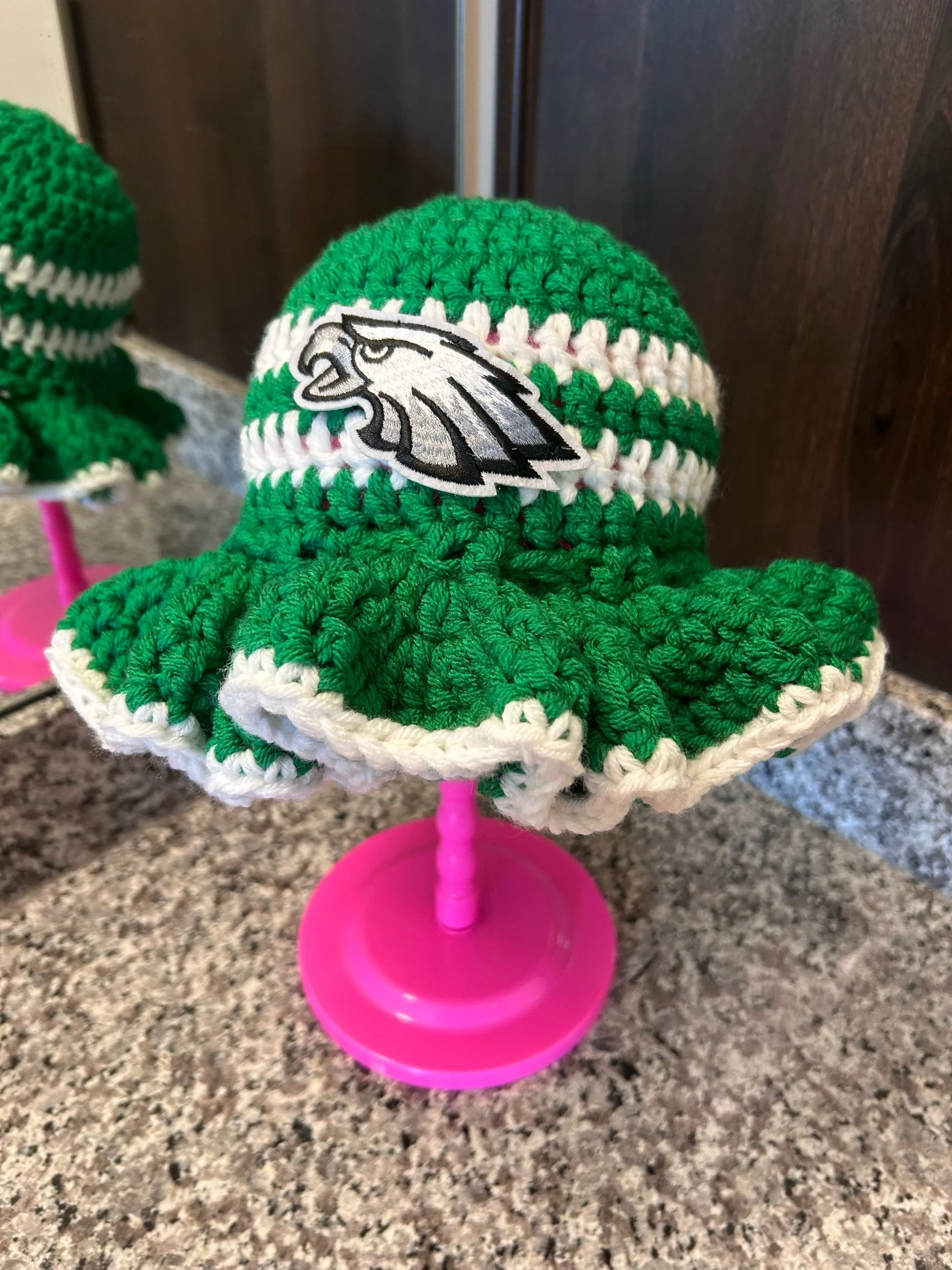 A handmade crocheted eagle logo cap in green, white, and black, displayed on a pink stand on a kitchen countertop.
