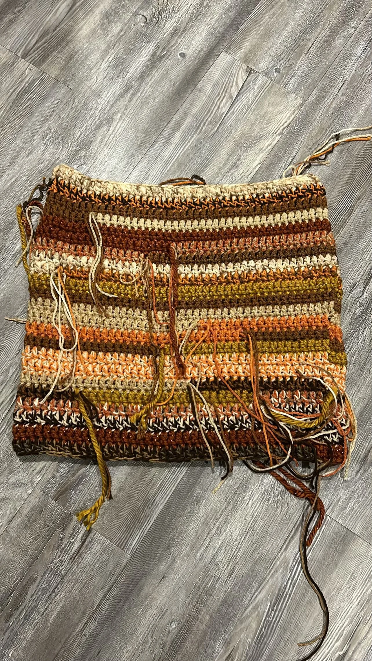 A partially finished knitted or crocheted fabric with multicolored yarn in shades of brown, orange, cream, and green, lying on a gray wooden floor. The fabric has loose yarn ends hanging from the edges.