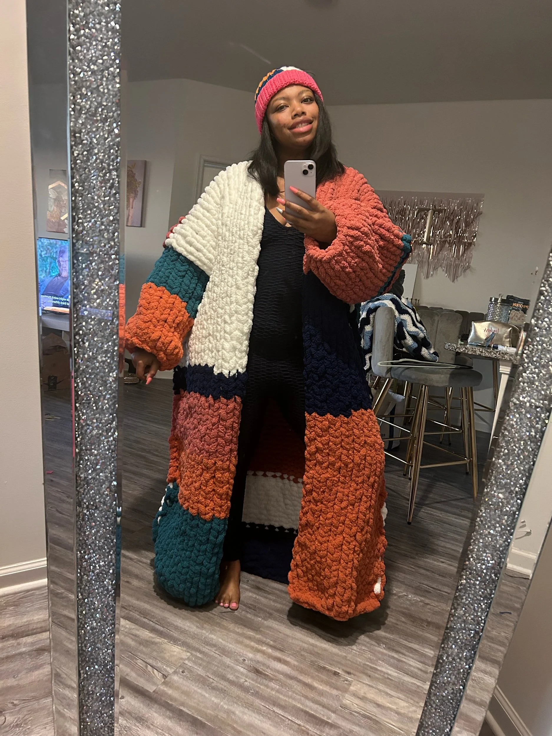 Woman taking a mirror selfie in a living room, wearing a colorful chunky knit sweater and a beanie.