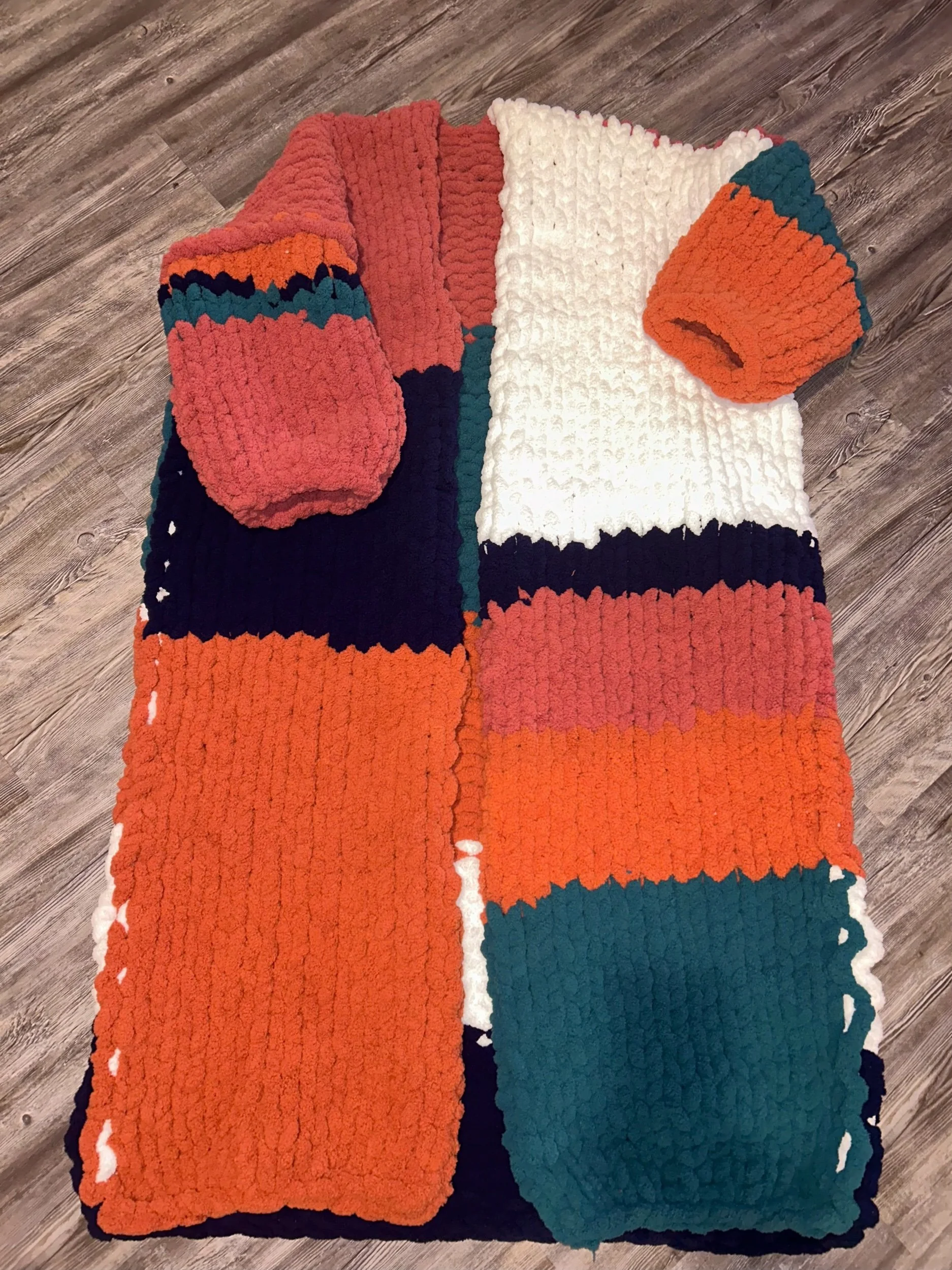 Colorful patchwork knitted blanket with white, orange, teal, navy, and coral sections, spread on a wooden floor.