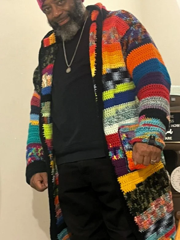 A man with a beard wearing a colorful, patchwork knitted coat over black clothing, standing indoors.