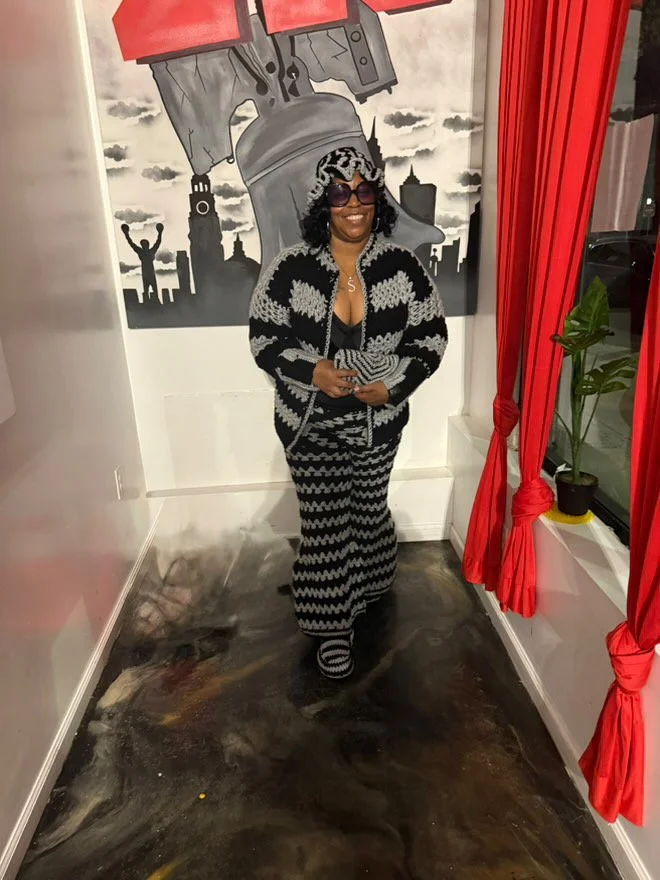 A woman wearing sunglasses, a black and white patterned outfit, and a headscarf, standing inside a room with a red curtain on the side, a potted plant, and a mural of the Statue of Liberty holding a large cannon in the background.