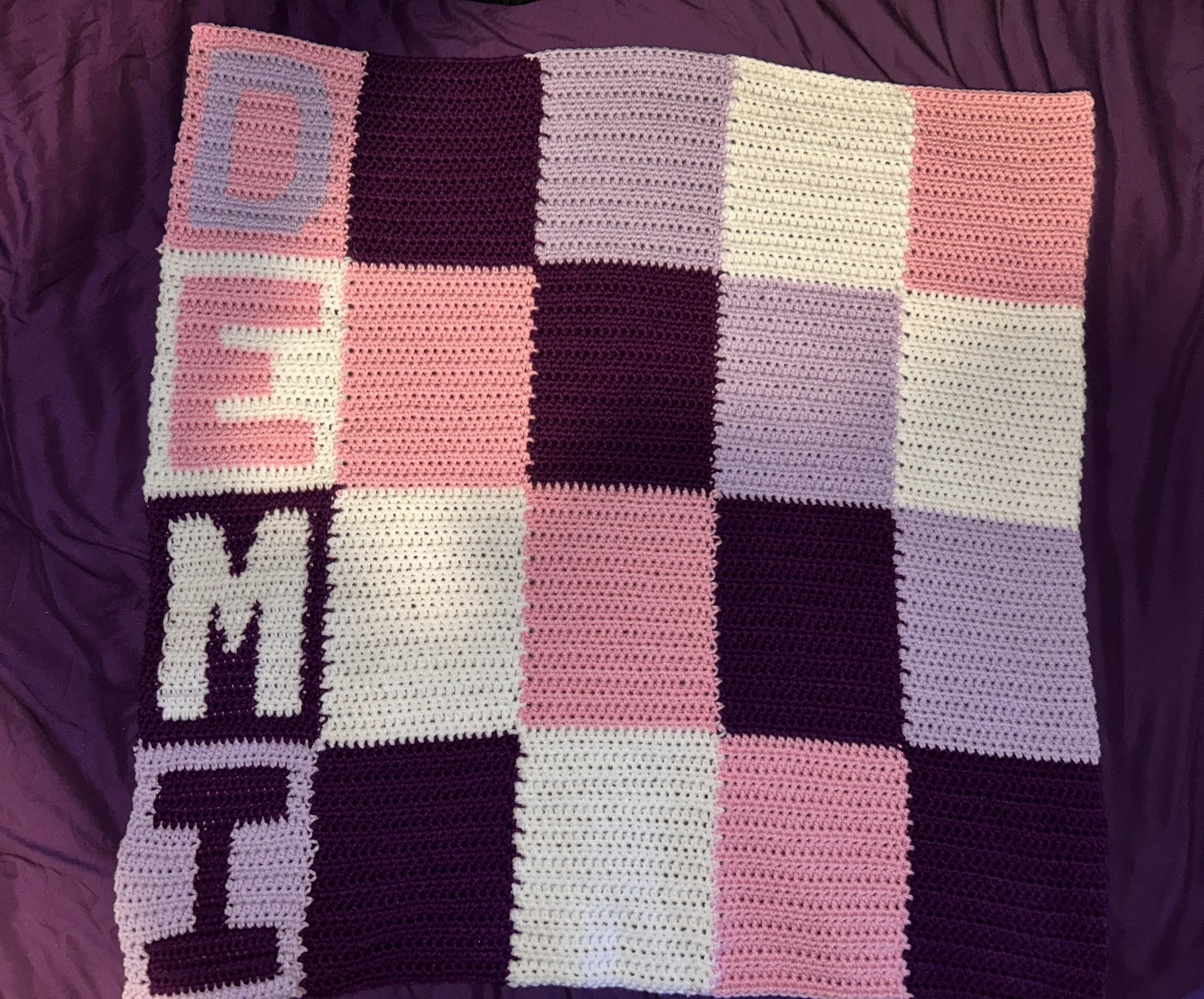 Handmade crocheted blanket with a checkered pattern of pink, purple, cream, and white squares, some with letters spelling 'LOVE' and 'MOM'.