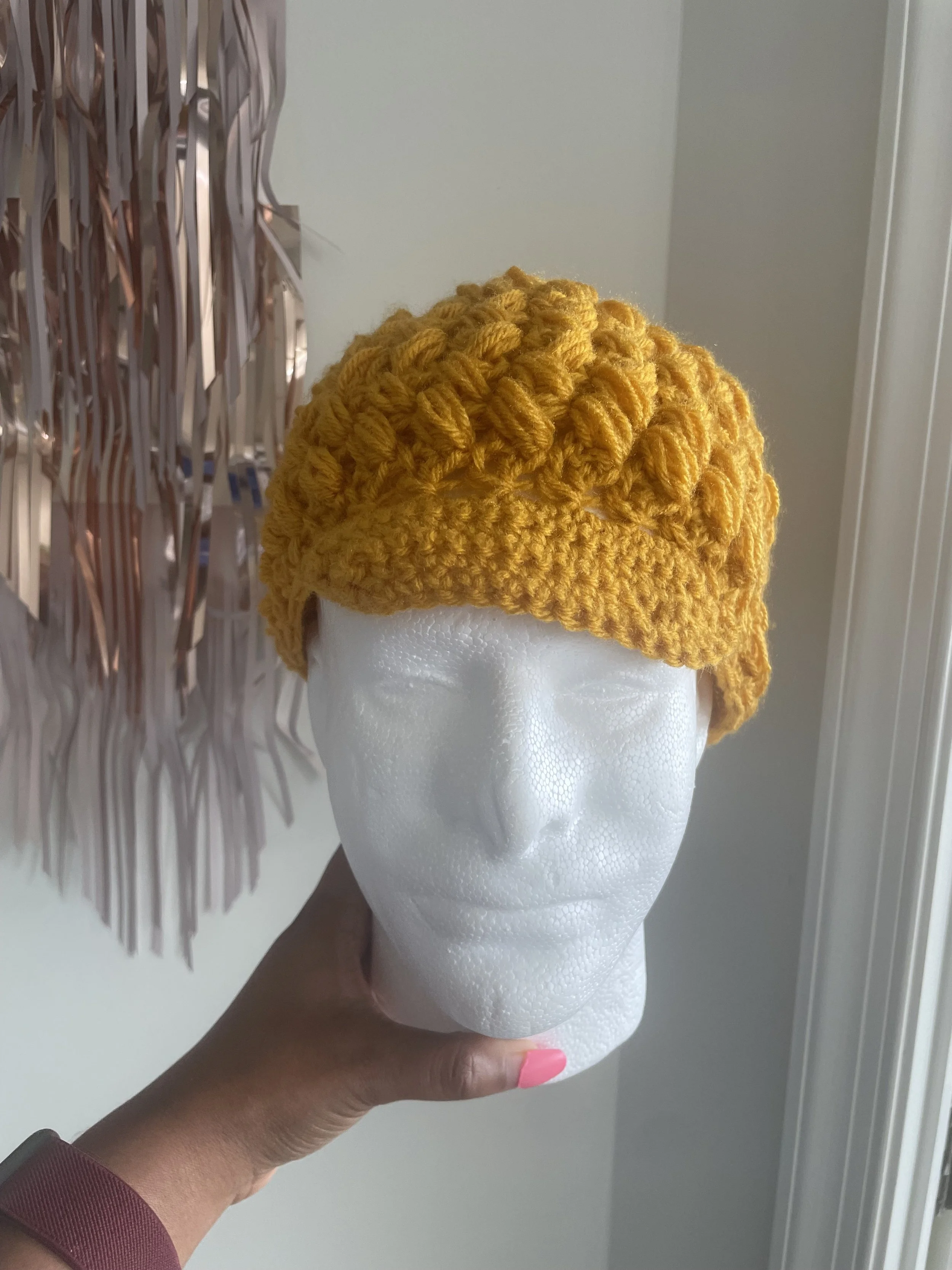 A mannequin head wearing a handmade yellow crochet hat with textured pattern, held by a person with pink painted nails, against a neutral background with decorative metallic wall art.