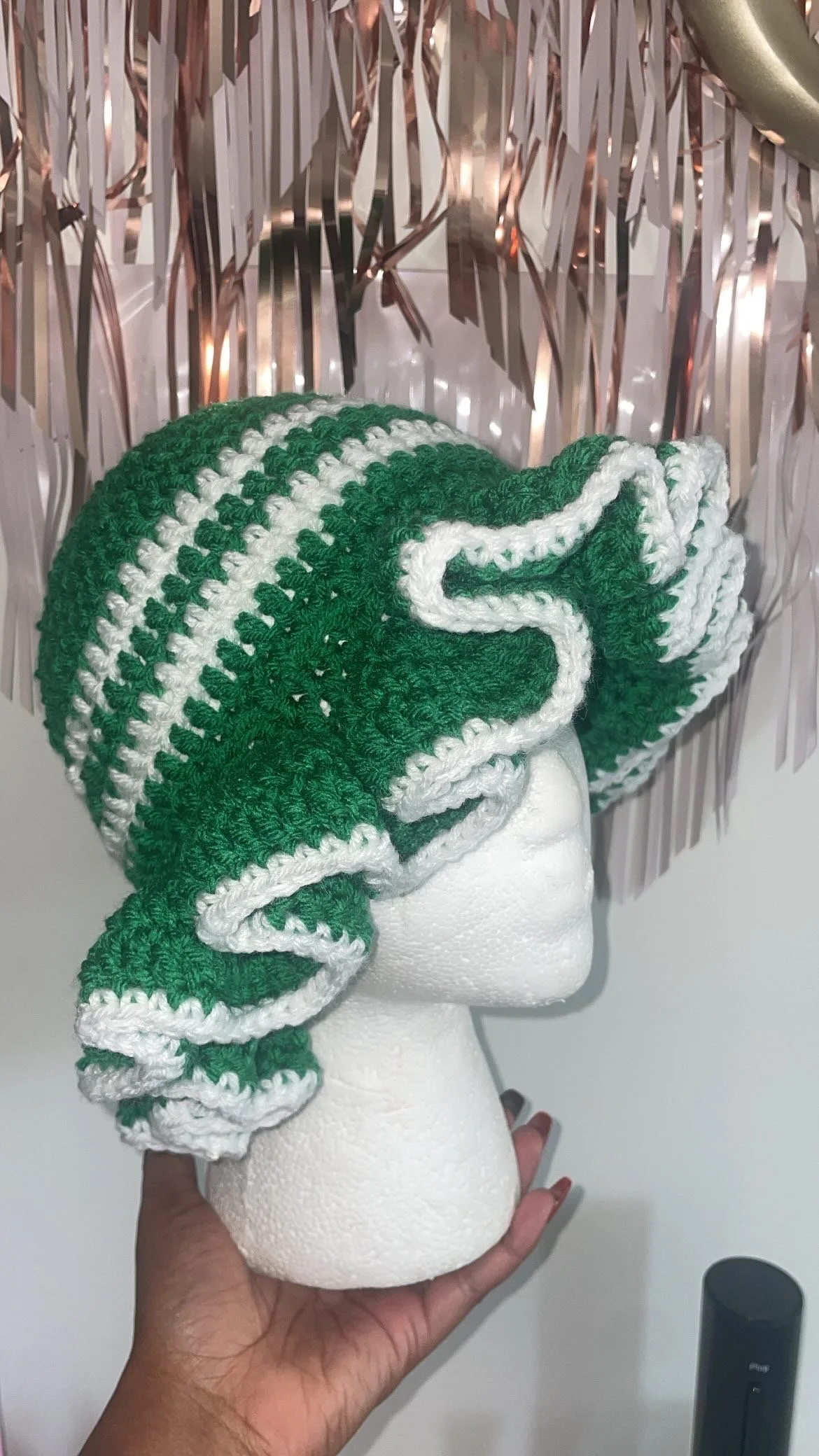 Hand holding a mannequin head with a crocheted green and white hat, in front of gold and rose gold tinsel decorations.