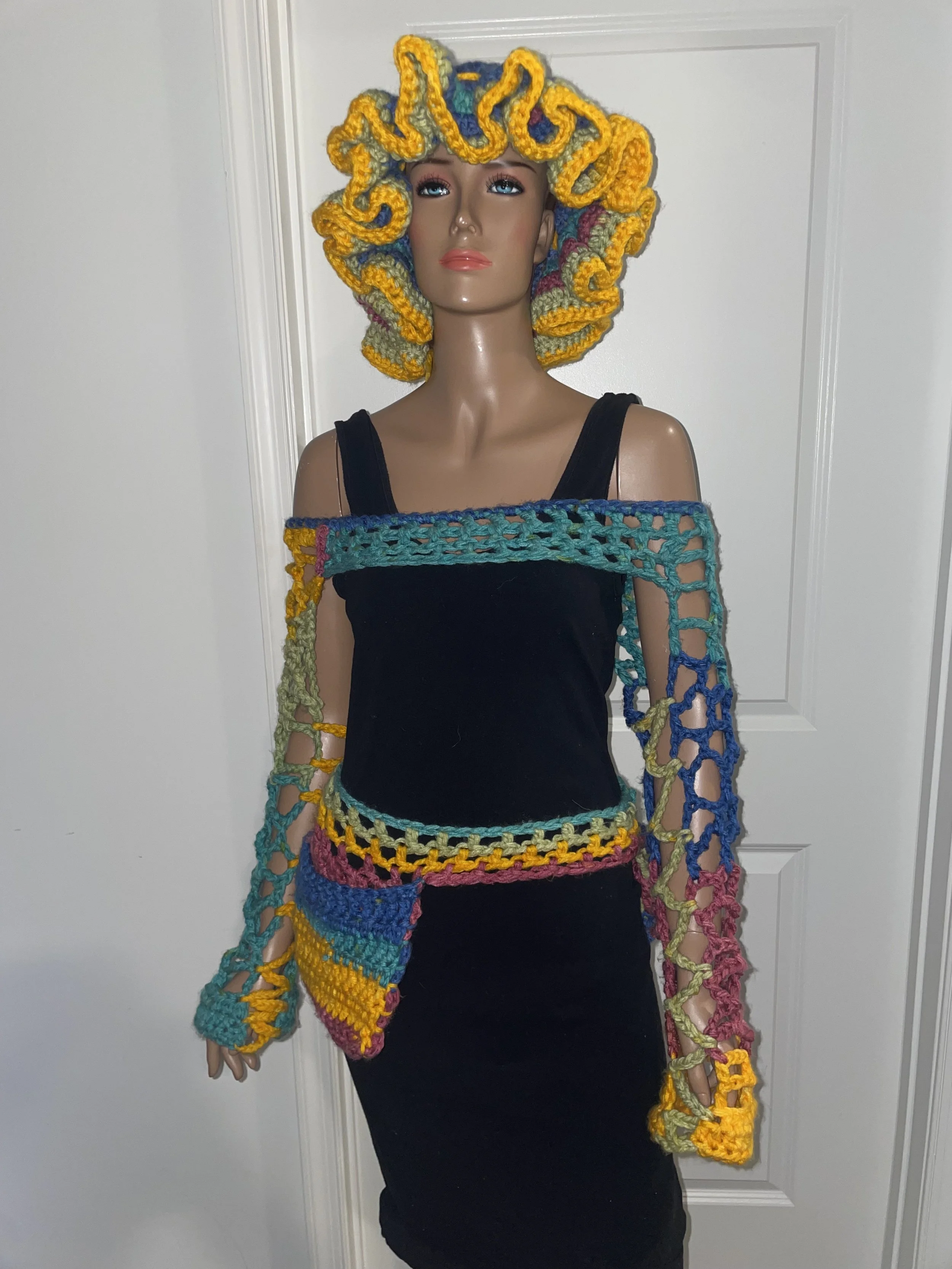 A mannequin dressed in a black tank top and black skirt, wearing a colorful handmade crochet sweater and a matching hat with yellow, blue, pink, and green yarn, standing in front of a white door.
