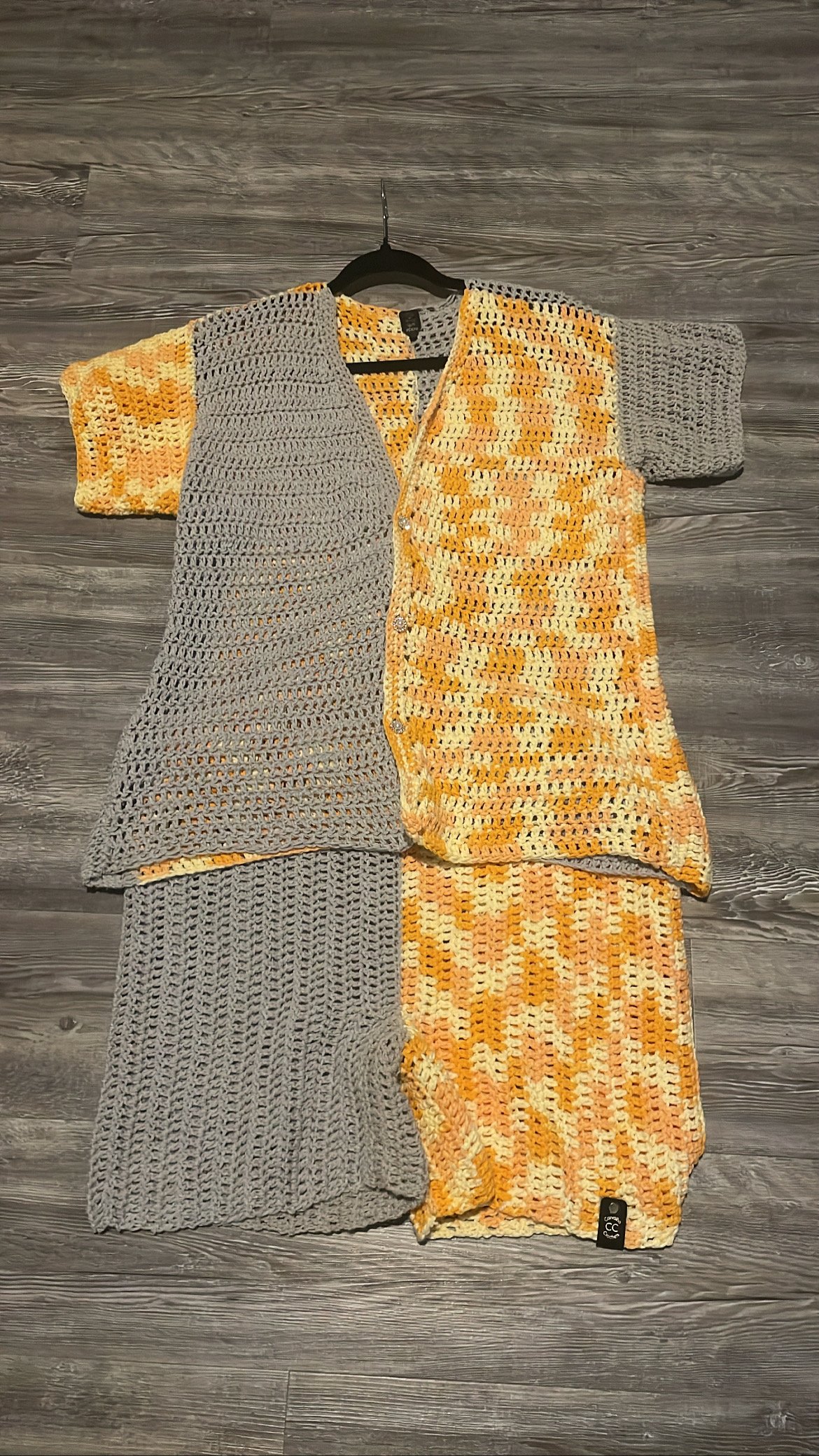 A handmade crochet dress with a patchwork design of grey and yellow/orange patterned sections, laid flat on a grey wooden floor.