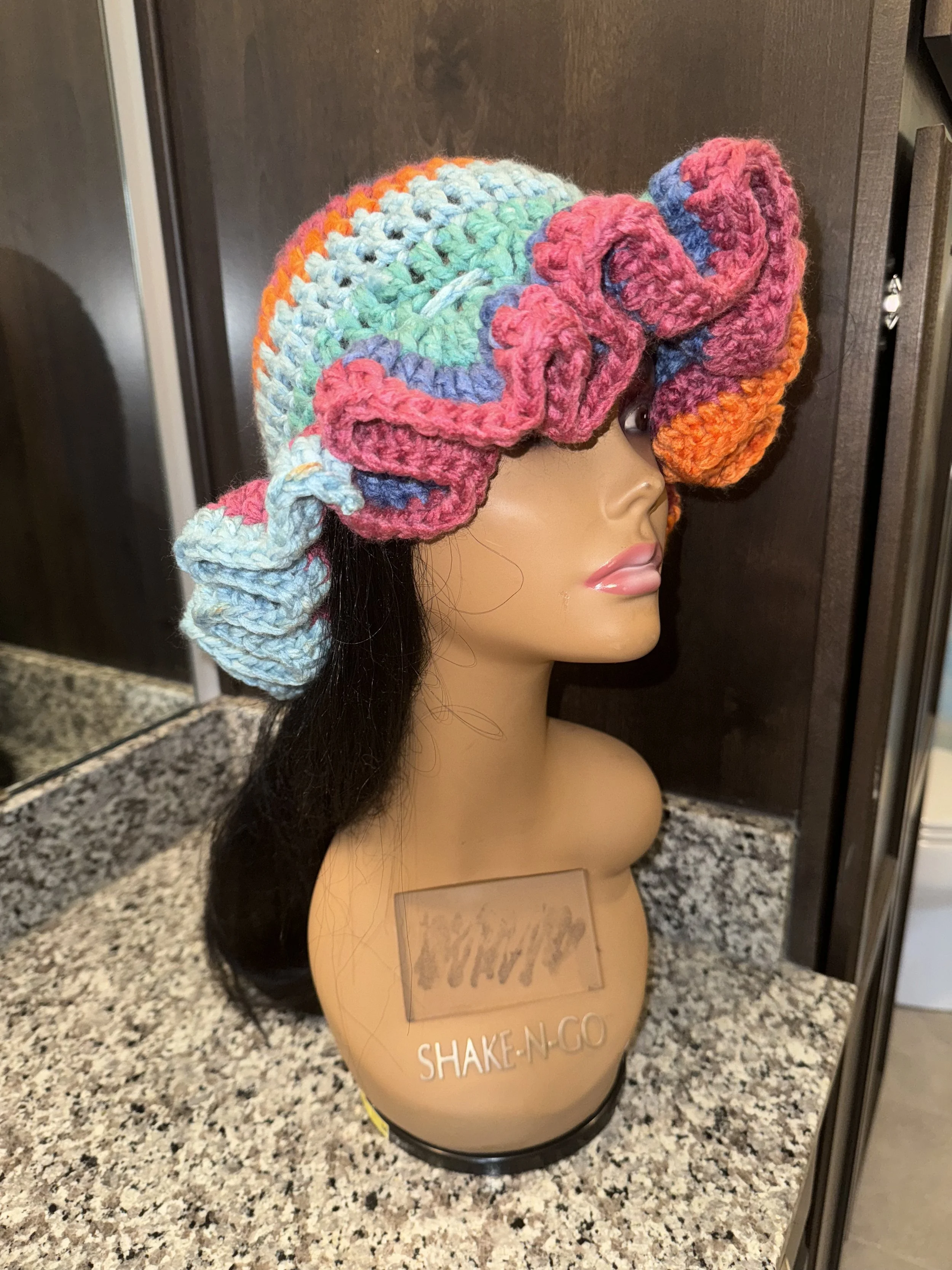 A mannequin head wearing a colorful, chunky knit hat with pink, blue, orange, and green yarns, placed on a speckled granite countertop with a dark wooden background.